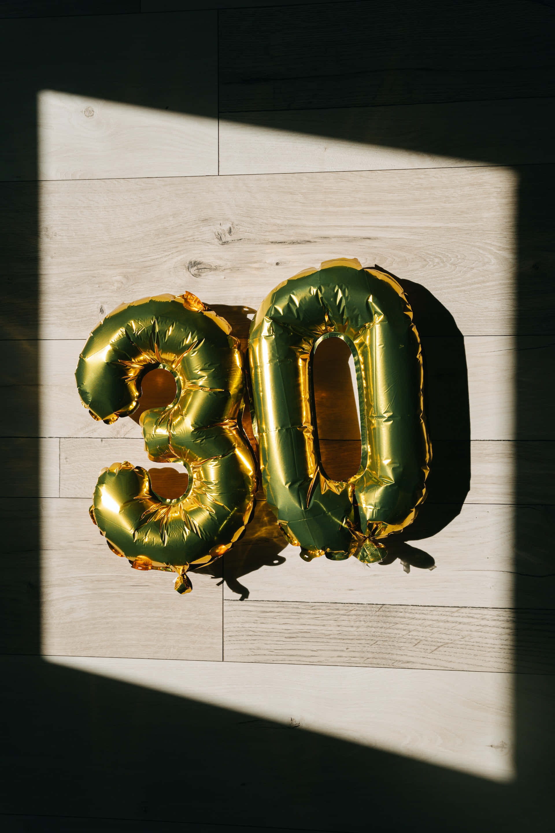 Number 桌布 壁紙 — 金色數字30箔氣球在淺色木地板上沐浴在強烈的陽光中，投射出戲劇性的陰影。