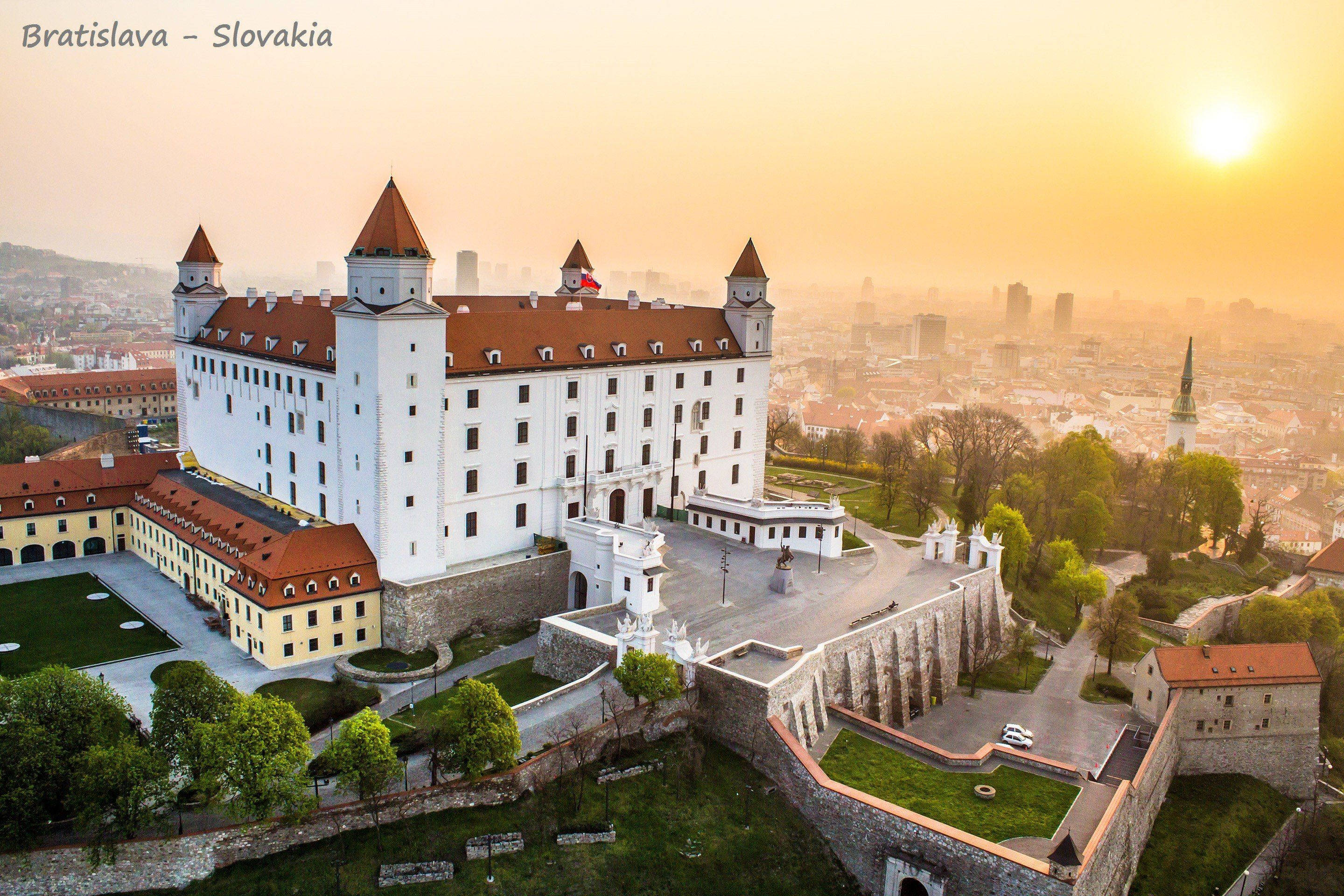Slovakia 桌布 壁紙 — 金色日出時從高空俯瞰布拉迪斯拉發城堡，溫暖的光線沐浴著白色牆壁和紅色屋頂，背景是朦朧的城市天際線。