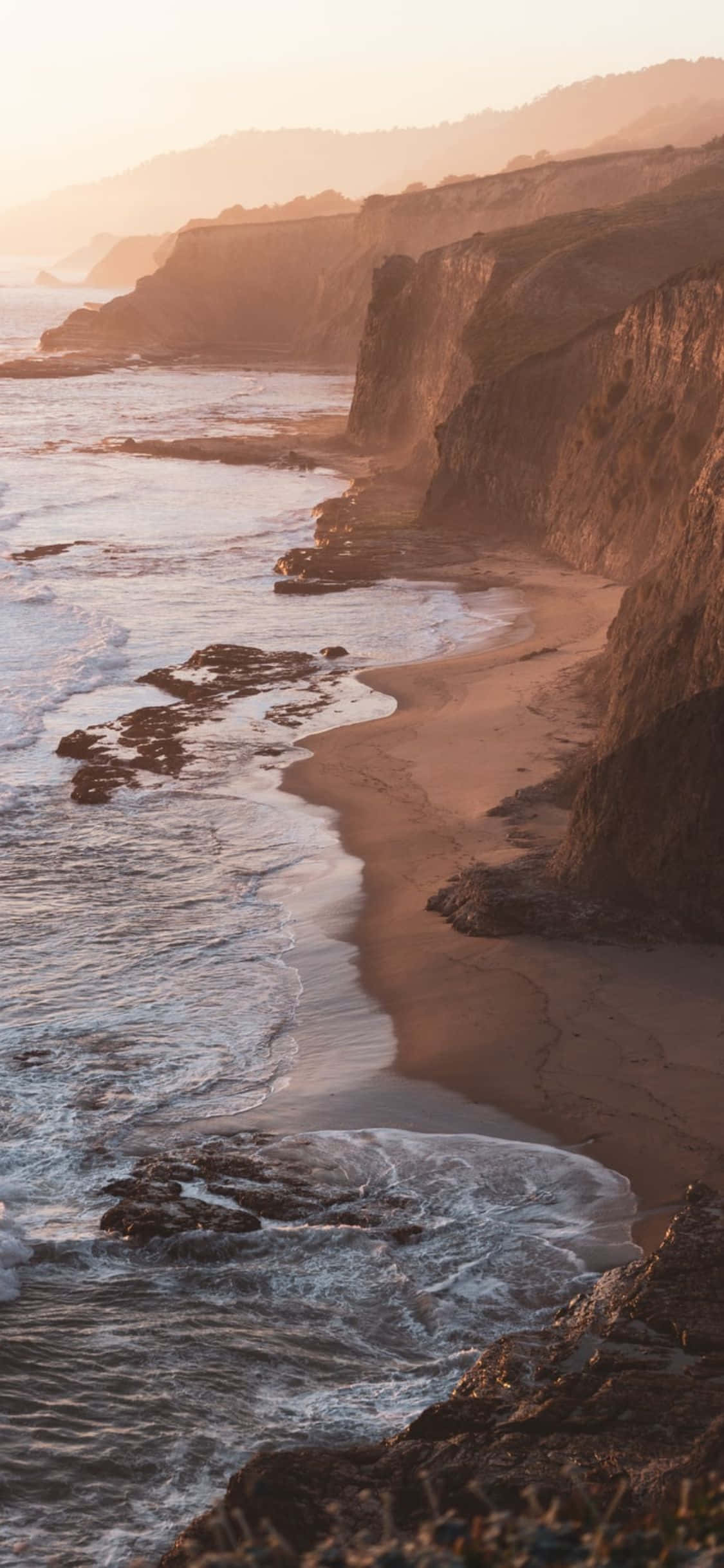 iPhone Xs California 桌布 壁紙 — 金色日出沐浴著崎嶇的海岸懸崖，海浪拍打著下方的沙灘。