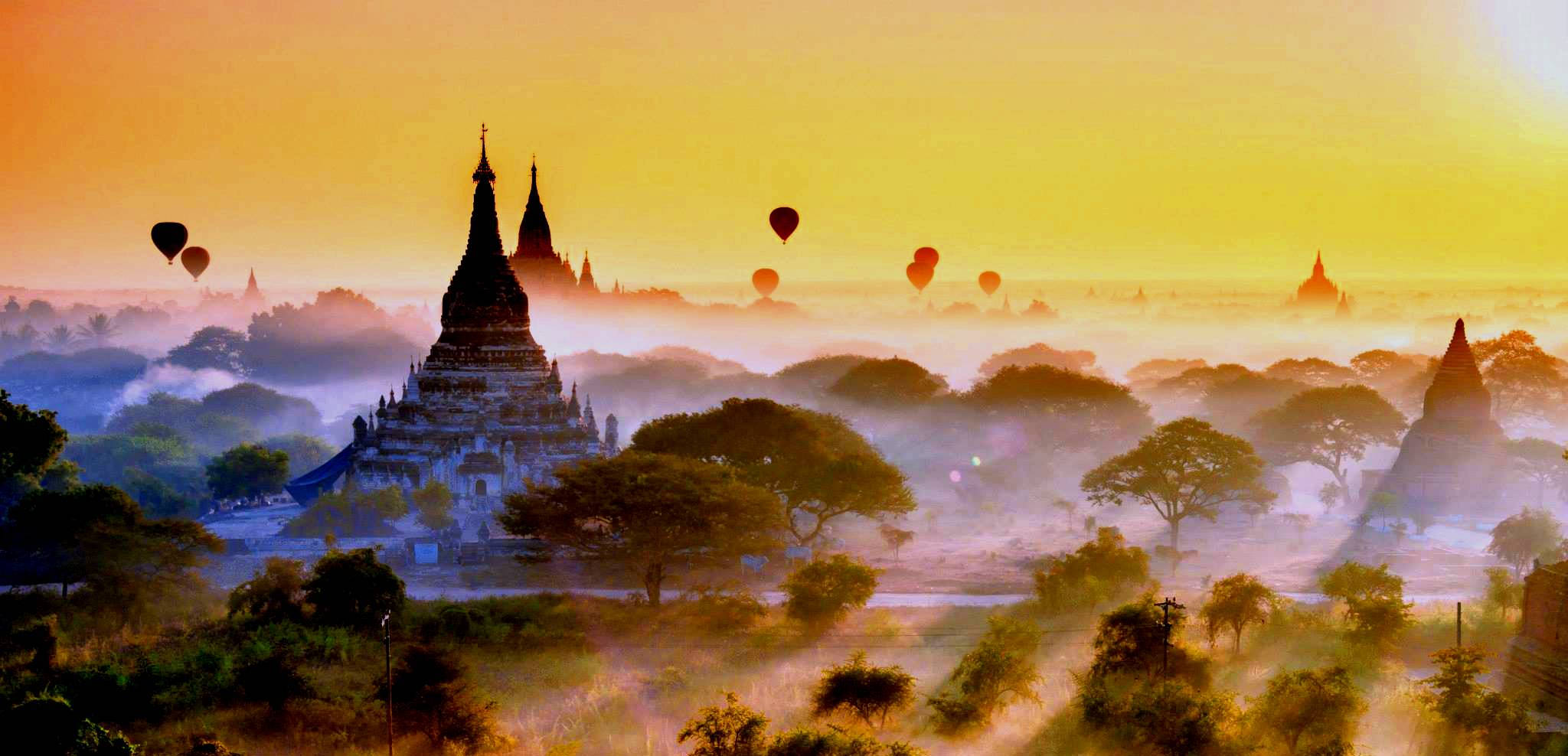 Yangon 桌布 壁紙 — 金色日出沐浴著緬甸古老佛塔的溫暖光芒，熱氣球在霧氣繚繞的樹木上方飄浮。