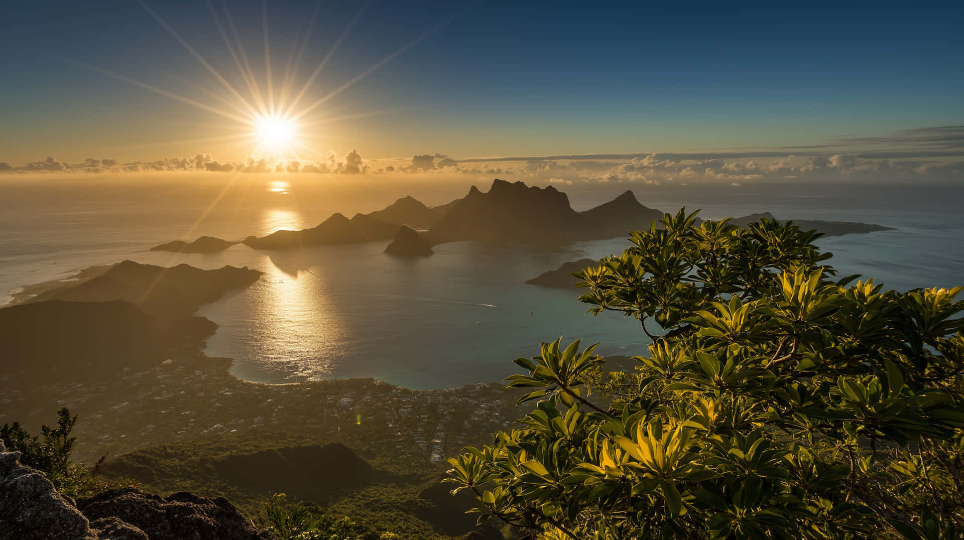 Sunrise Over Tropical Mountains and Shimmering Ocean 壁紙 — 金色日出照耀著熱帶海灣，有鋸齒狀的火山島和前景茂密的綠色樹葉。