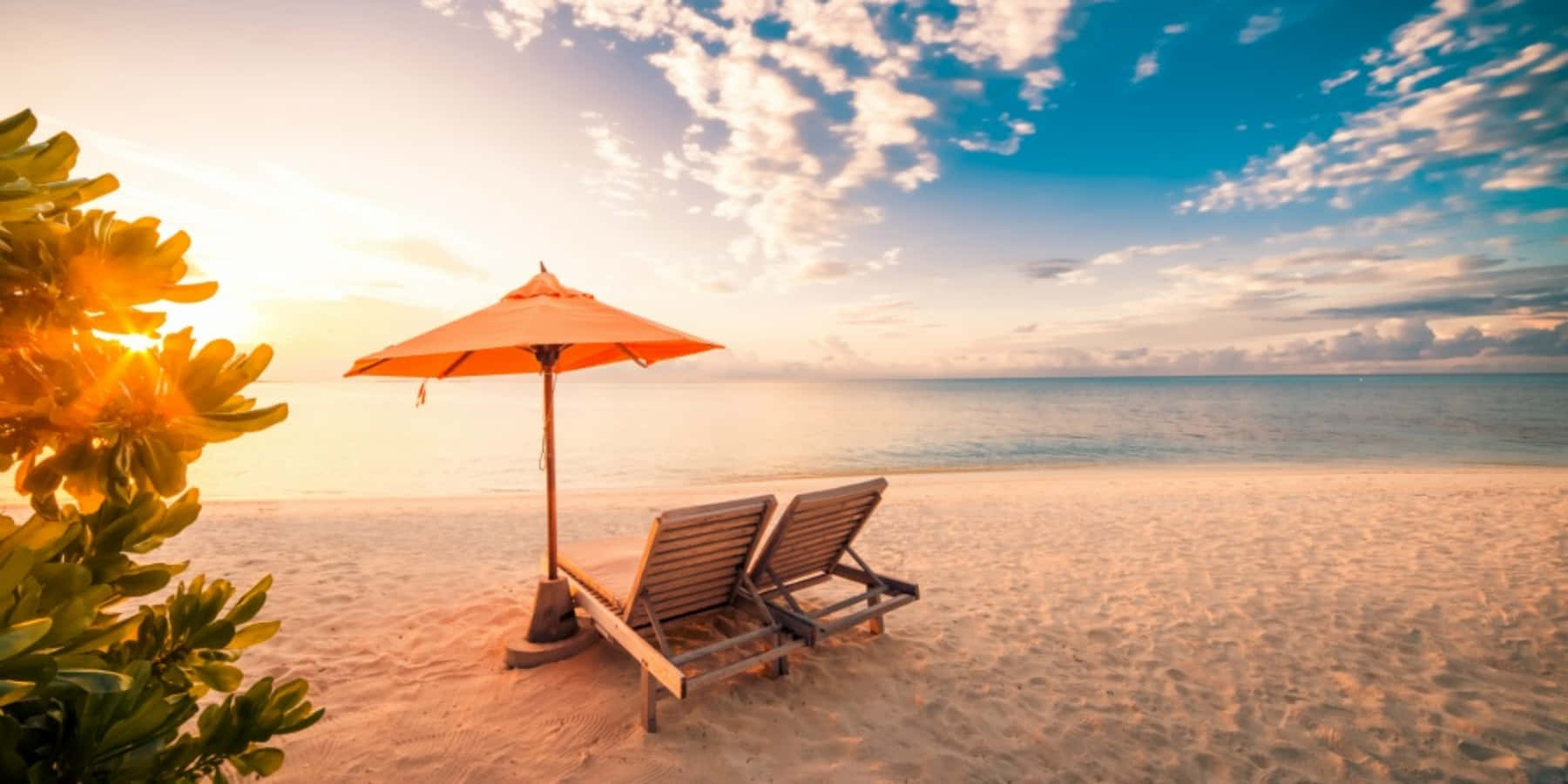 Vacation 桌布 壁紙 — 金色日落時分，沙灘上橙色遮陽傘下的兩張木製躺椅，前景有棕櫚葉。