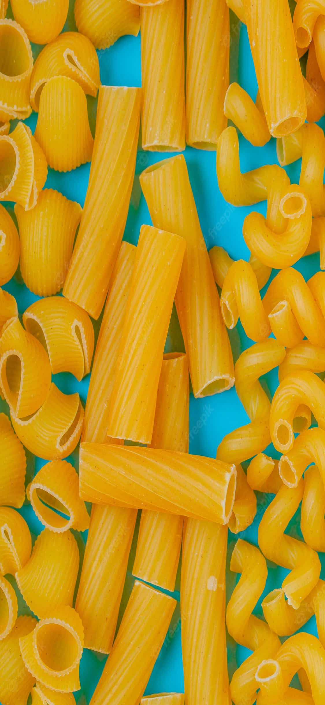 iPhone X Pasta 桌布 壁紙 — 金色未煮熟的義大利麵管和貝殼散落在充滿活力的藍色背景上，以俯視平鋪方式呈現。