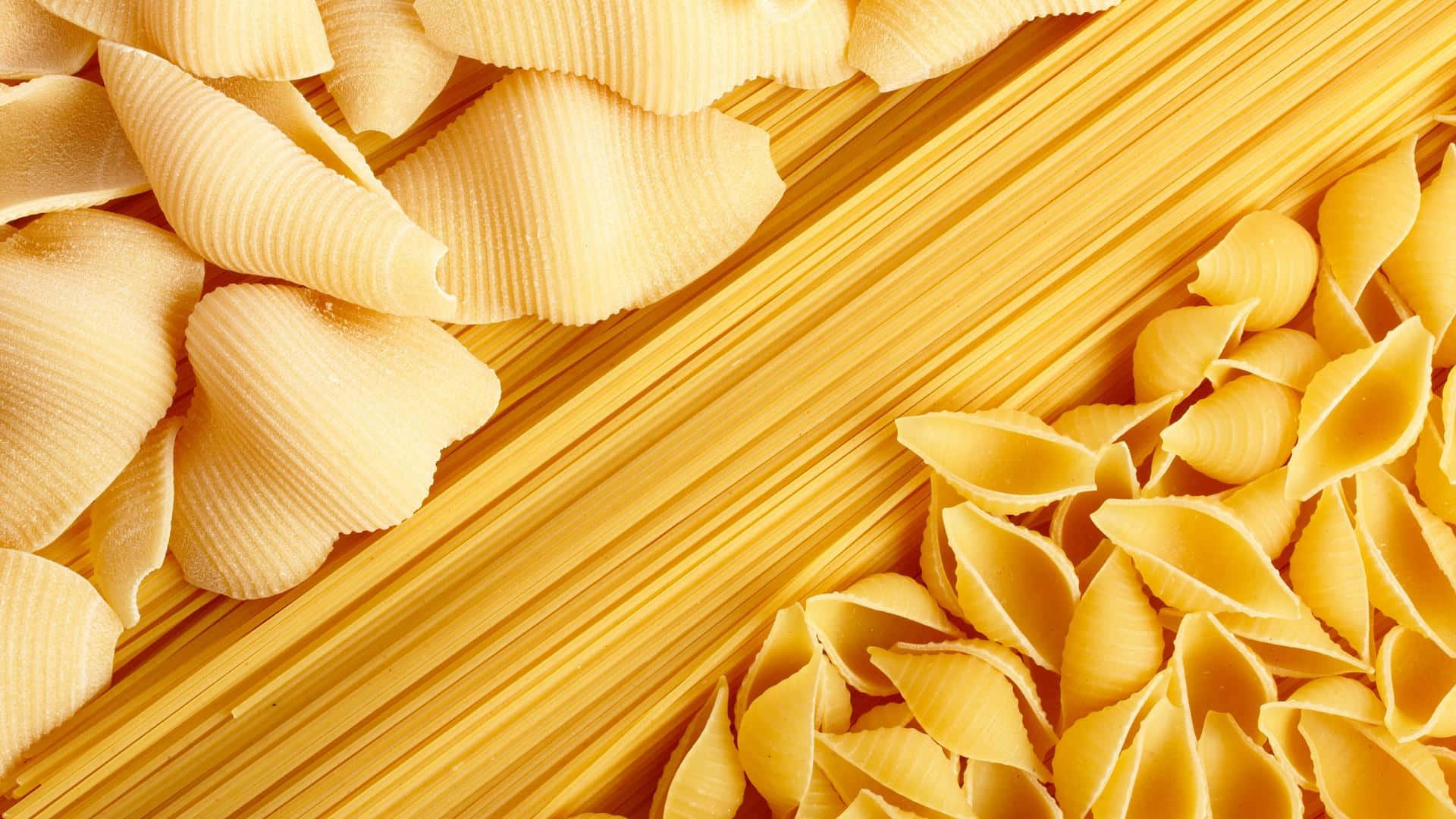Pasta 桌布 壁紙 — 金色未煮熟的義大利麵貝殼和義大利麵條以對角線排列，形成充滿活力的特寫構圖。