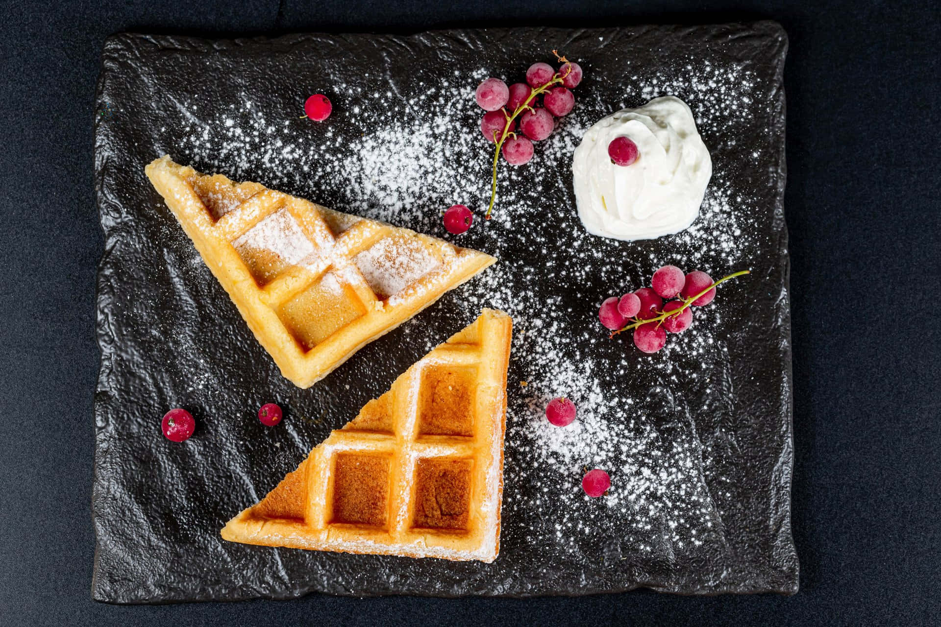 Waffle 桌布 壁紙 — 金色格子鬆餅三角形撒上糖粉，並以紅葡萄和鮮奶油點綴，放在深色石板盤上。