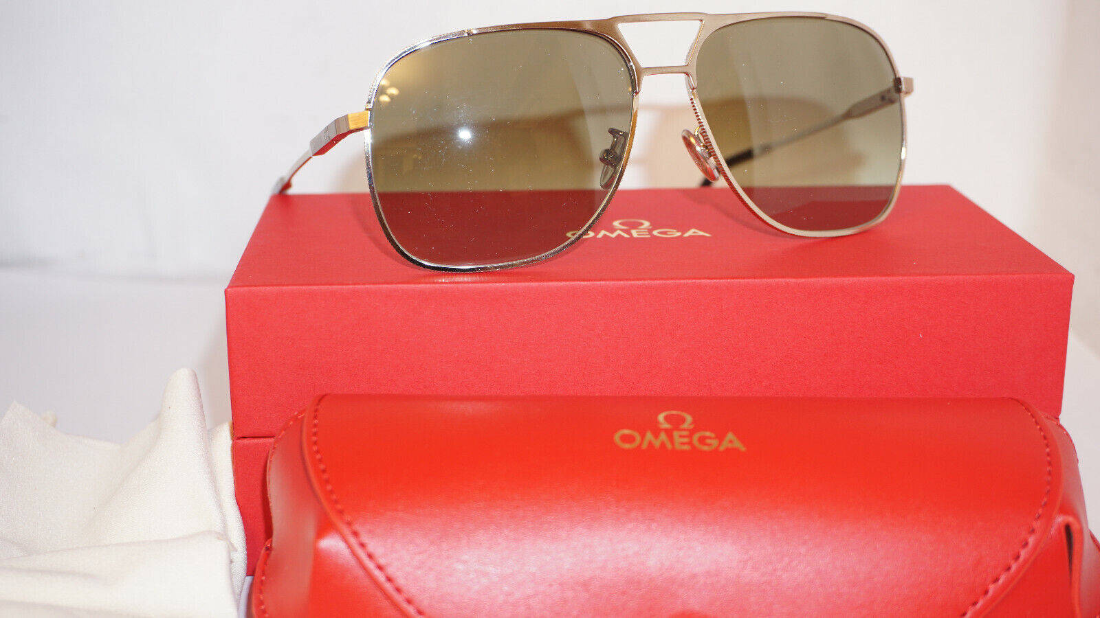 Sunglasses 桌布 壁紙 — 金色框飛行員太陽眼鏡放在白色背景上的紅色Omega盒子和相配的保護盒上。