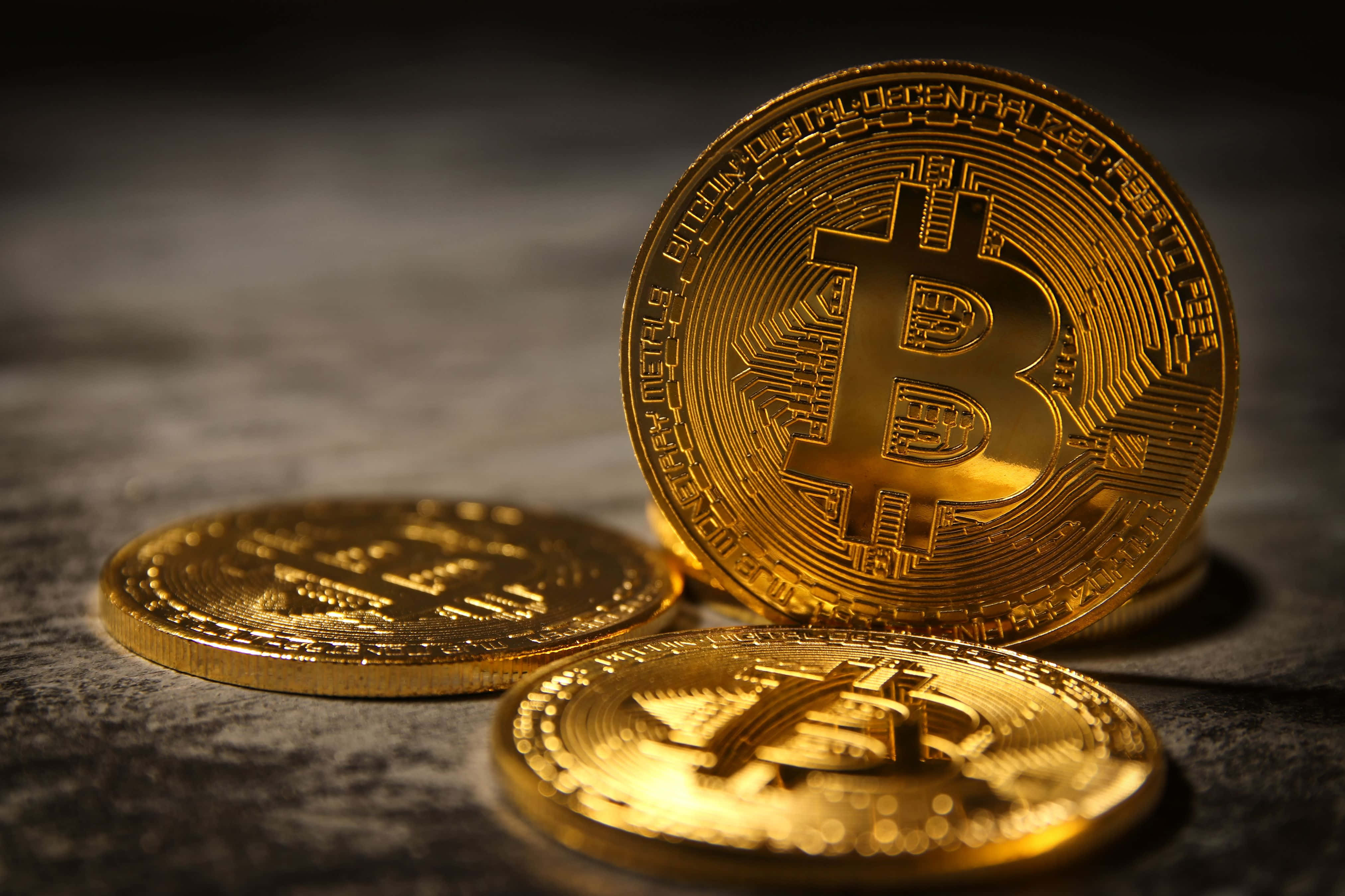 4K Bitcoin 桌布 壁紙 — 金色比特幣在戲劇性燈光下，於淺景深的深色紋理表面上閃閃發光。