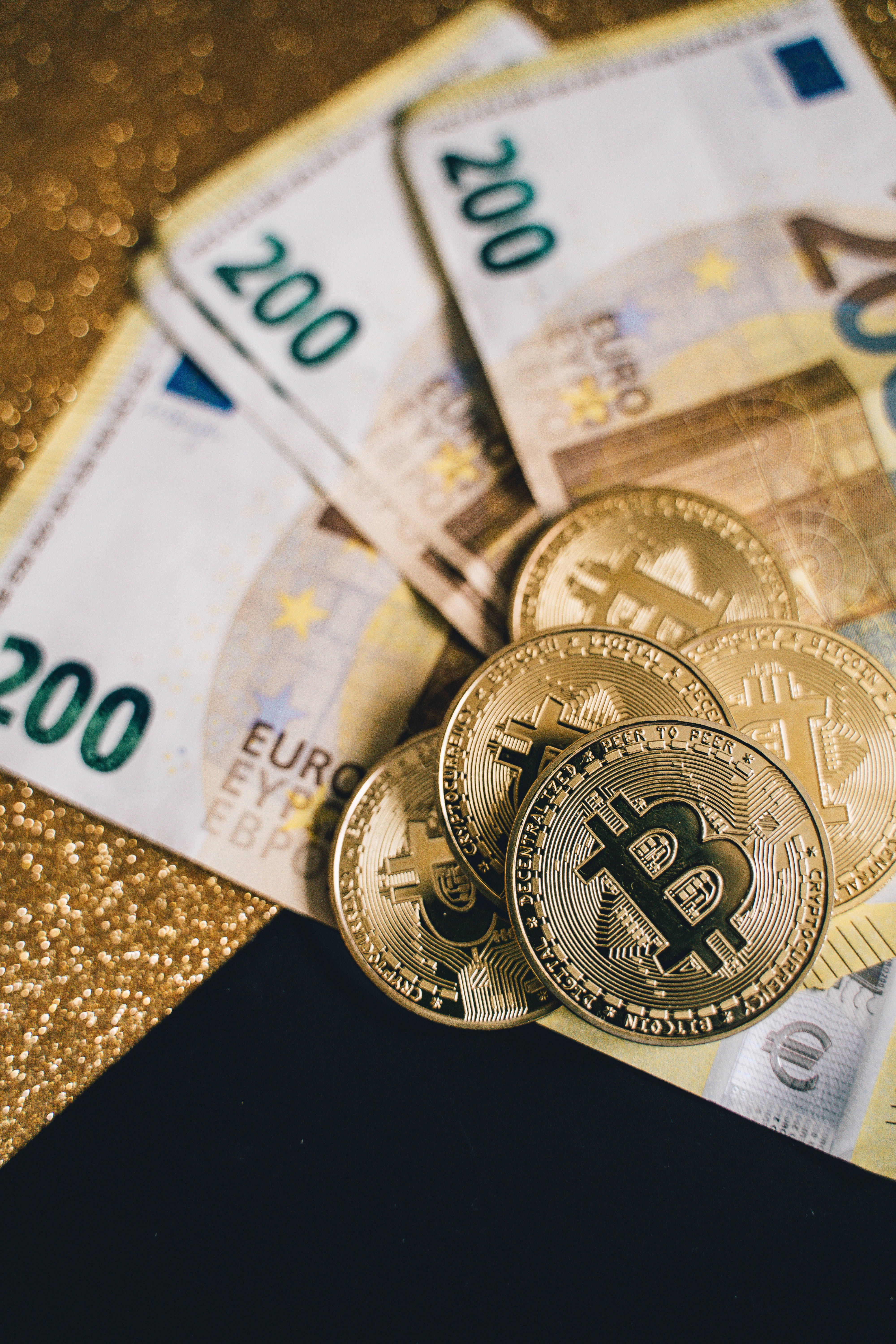 金錢 桌布 壁紙 — 金色比特幣硬幣放在散落的200歐元紙幣上，表面閃爍金黑光澤。