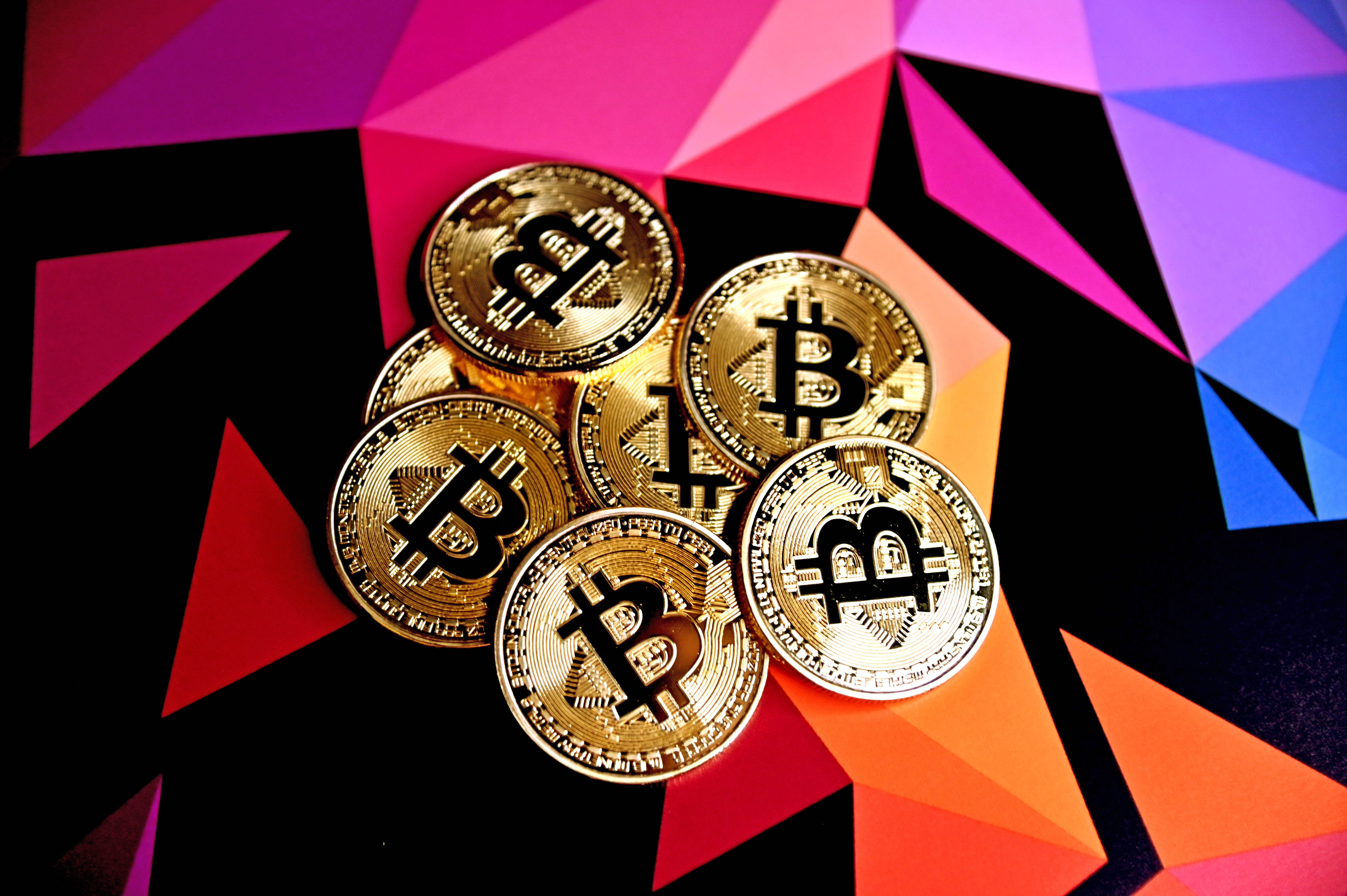 比特幣 桌布 壁紙 — 金色比特幣硬幣放置在由粉紅色、紫色、藍色和橙色三角形組成的鮮豔幾何背景上。