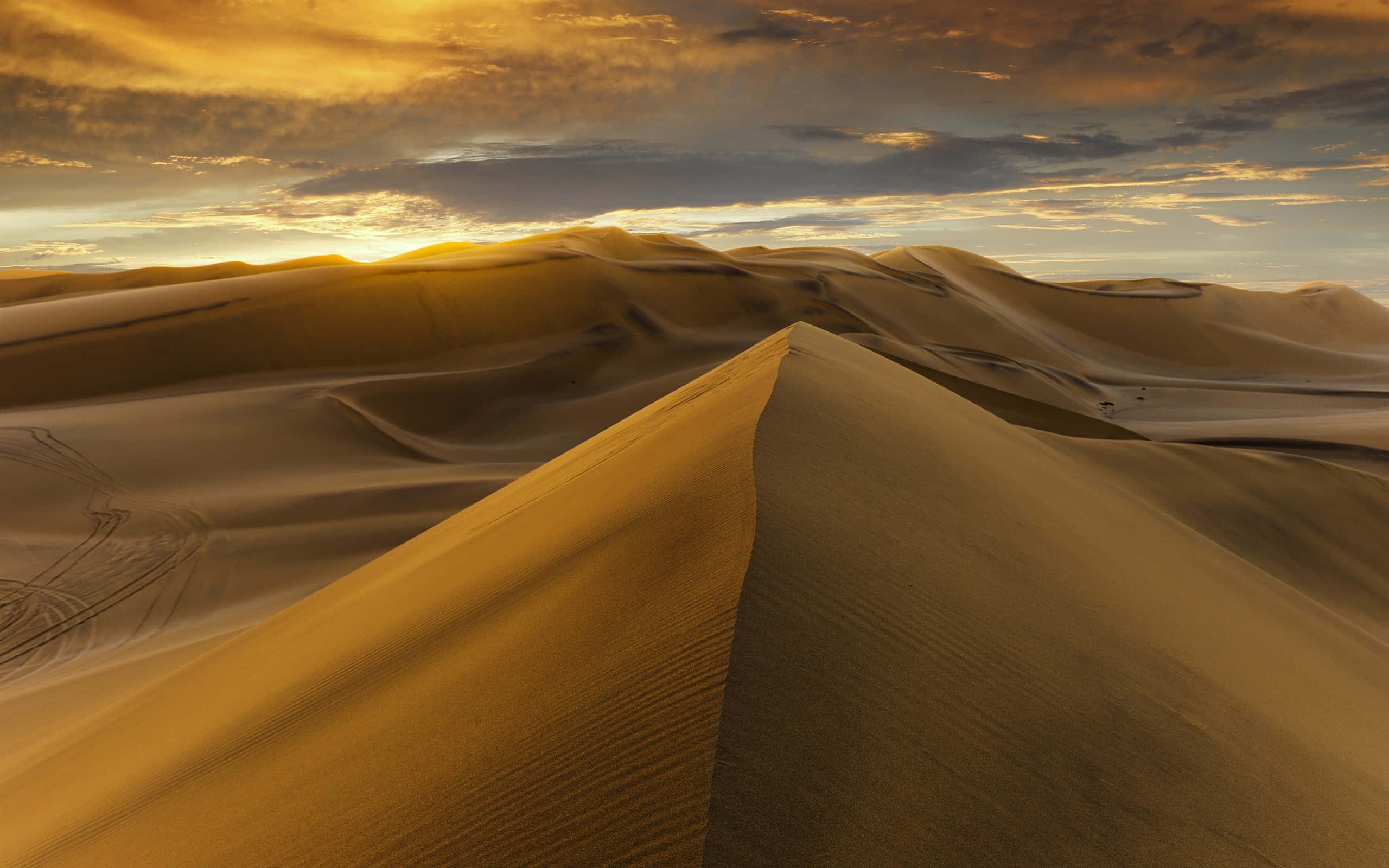Sand Dunes 桌布 壁紙 — 金色沙丘在壯麗的日落天空下起伏，天空中有橙色和藍色的雲彩條紋。