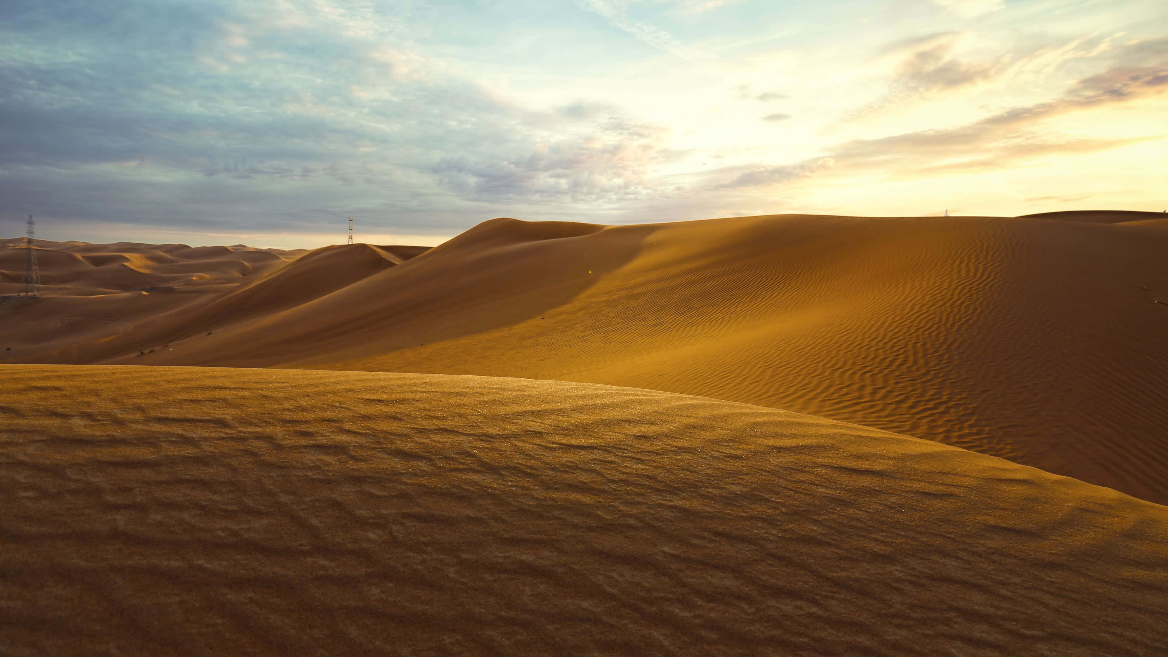 Sand Dunes 桌布 壁紙 — 金色沙丘在柔和的日落天空下起伏，有零星雲朵和遠處的電線。