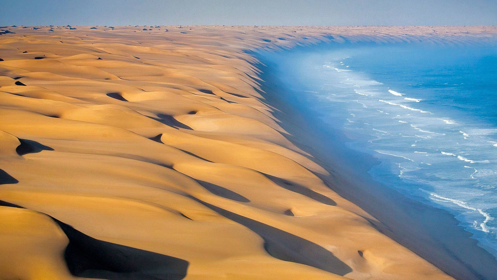 Namibia 桌布 壁紙 — 金色沙漠沙丘在清澈藍天下，在超現實的海岸景觀中與碧綠海洋波浪相遇。