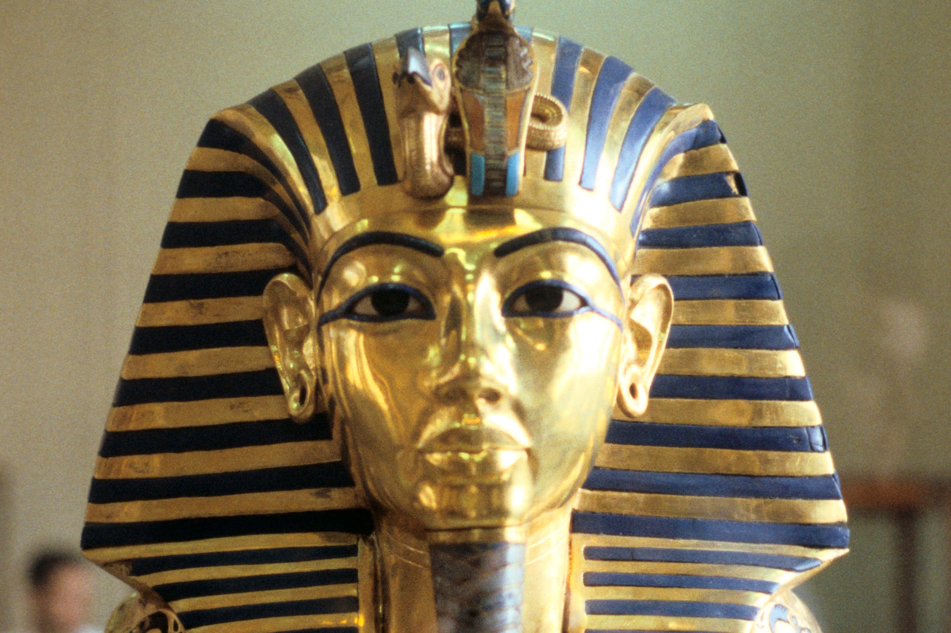 Ancient Egypt 桌布 壁紙 — 金色法老王面具帶有藍色條紋和眼鏡蛇標誌，在柔和的博物館燈光下閃閃發光。