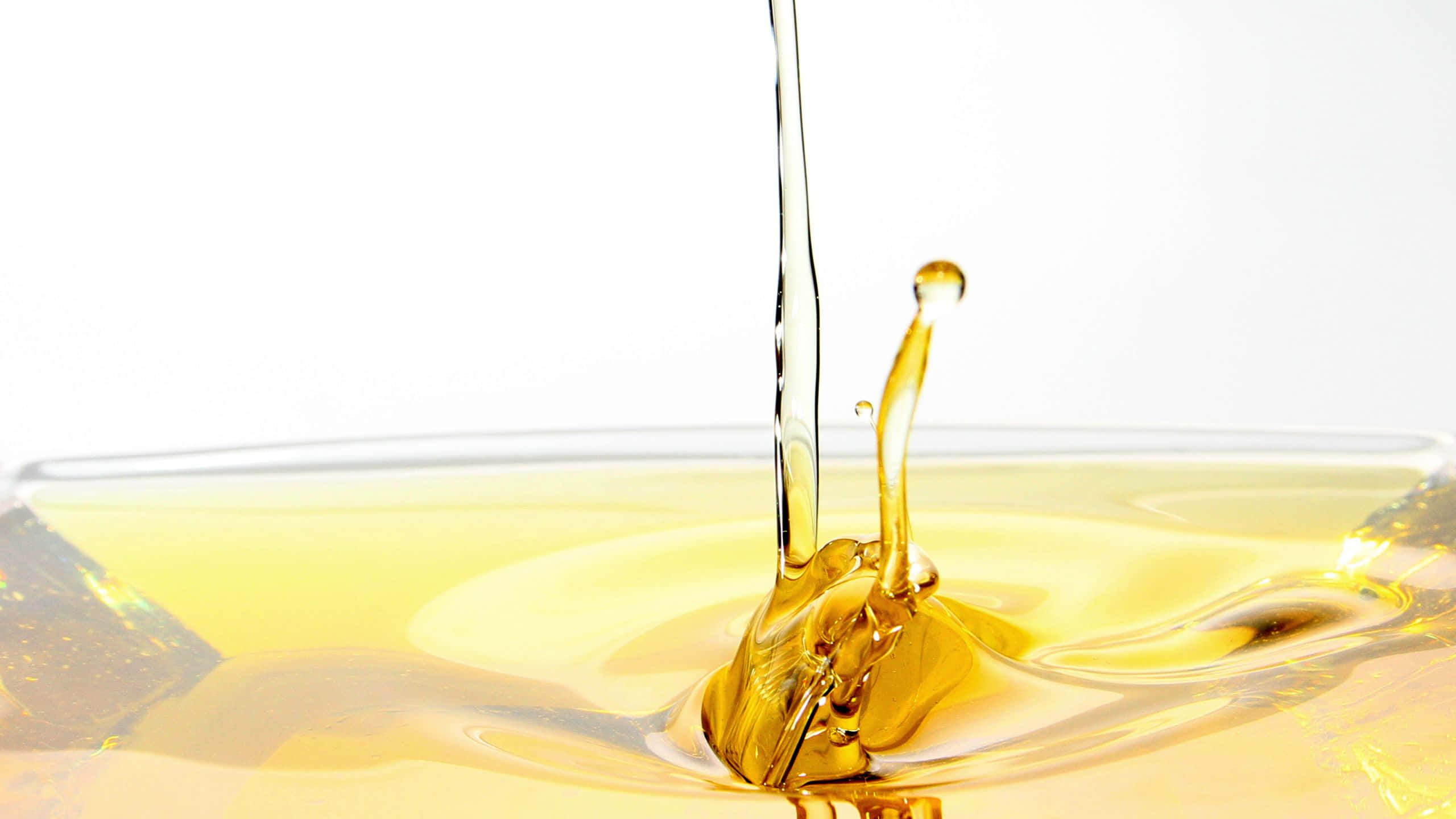 Oil 桌布 壁紙 — 金色液體流濺入玻璃碗中，在純白背景上形成動態漣漪和飛濺的水滴。