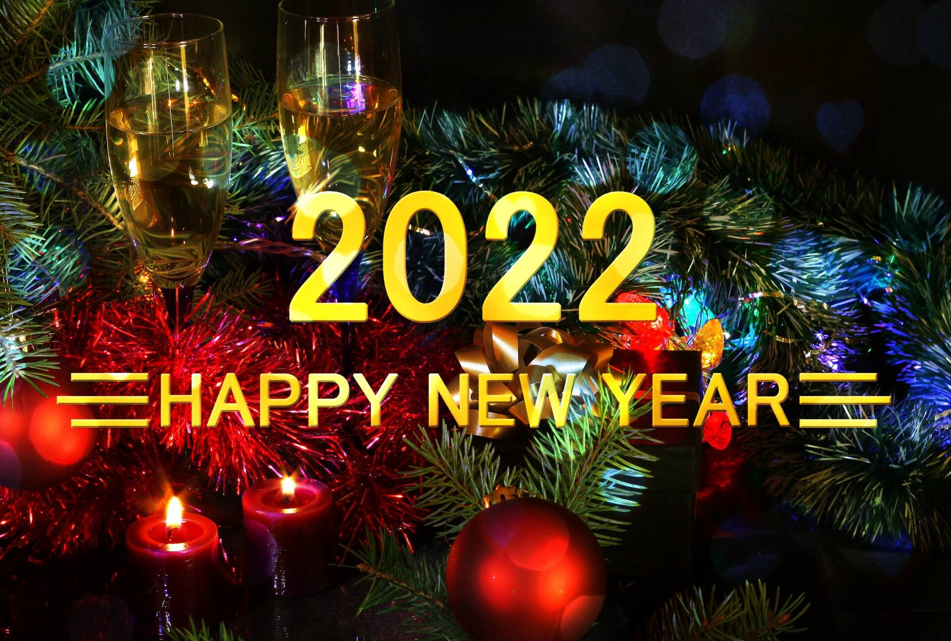 New 桌布 壁紙 — 金色的 '2022 HAPPY NEW YEAR' 文字在黑暗的散景背景上，在節日松樹枝、蠟燭和香檳杯上方發光。