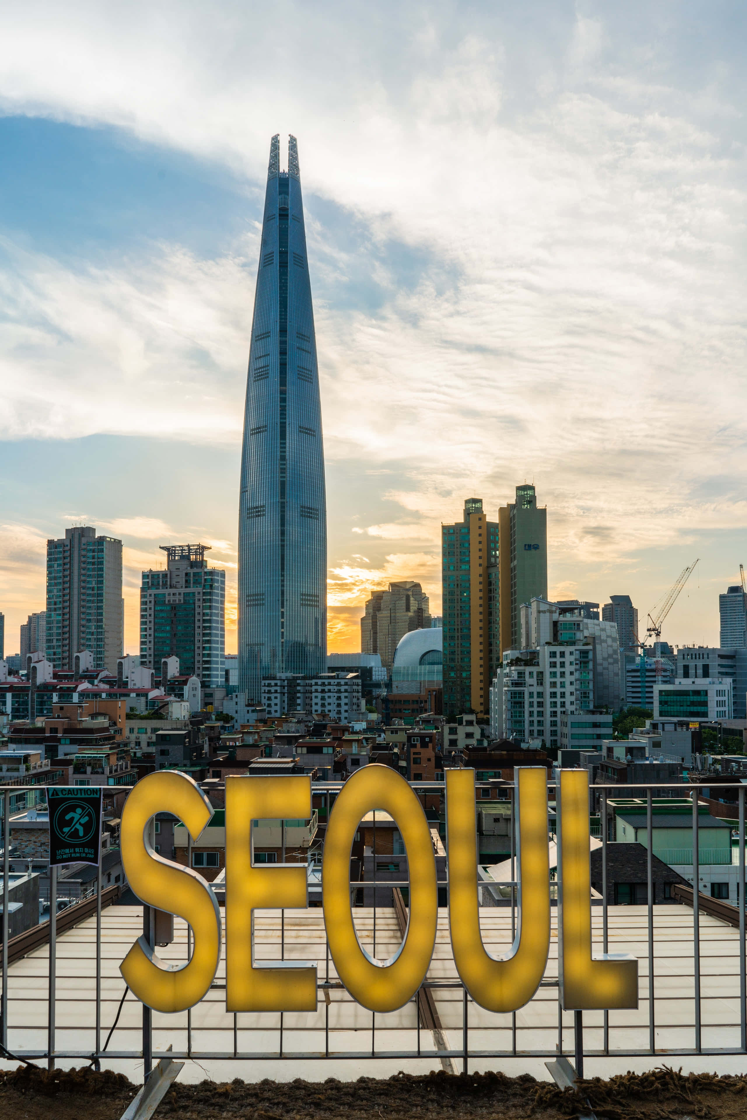 南韓 桌布 壁紙 — 金色的「SEOUL」字母在日落背景下發光，樂天世界塔在密集的城市天際線上刺穿天空。
