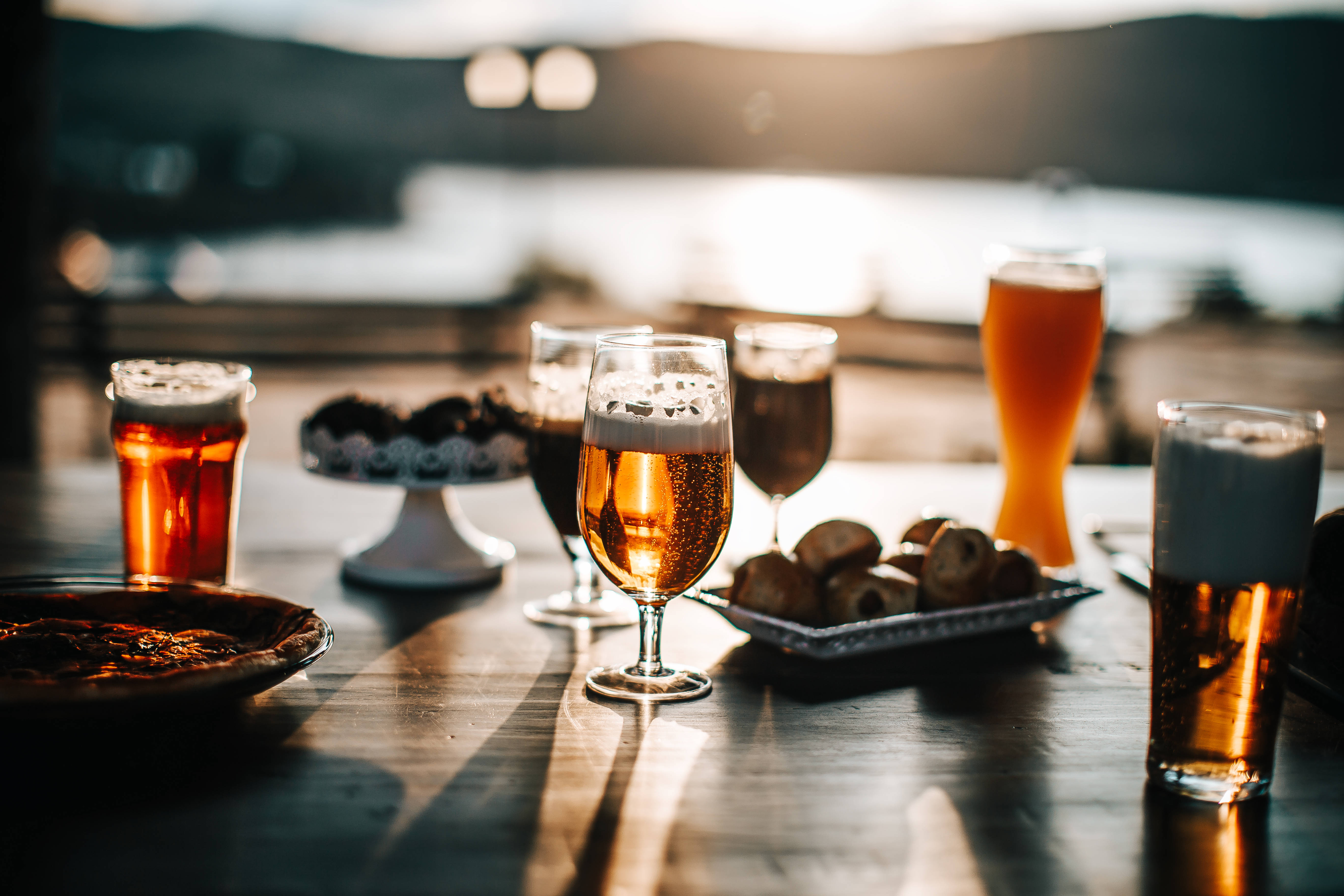 Beer 桌布 壁紙 — 金色的啤酒杯和小吃在木桌上溫暖地閃耀，夕陽的光線將海濱場景沐浴在柔和的琥珀色調中。