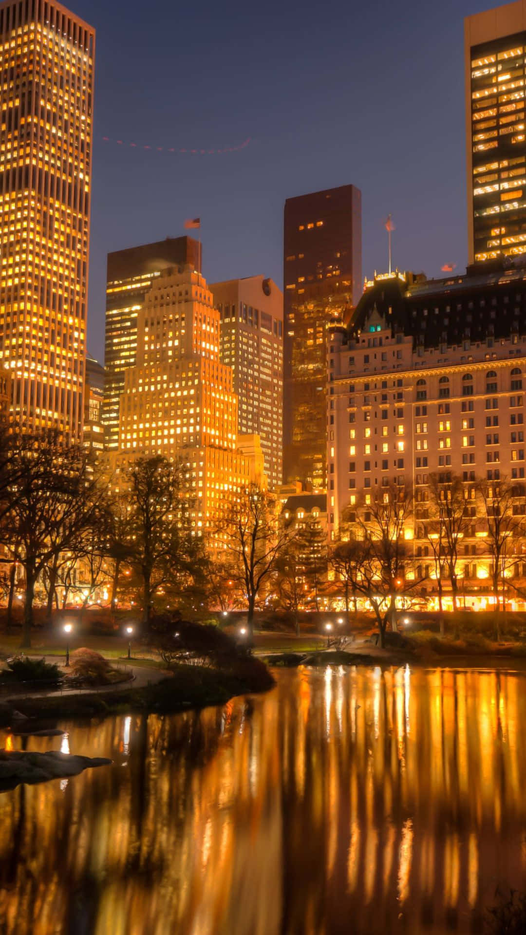 Android Ny City 桌布 壁紙 — 金色的城市燈光在黃昏時分，在高聳的摩天大樓下的平靜池塘上溫暖地反射。