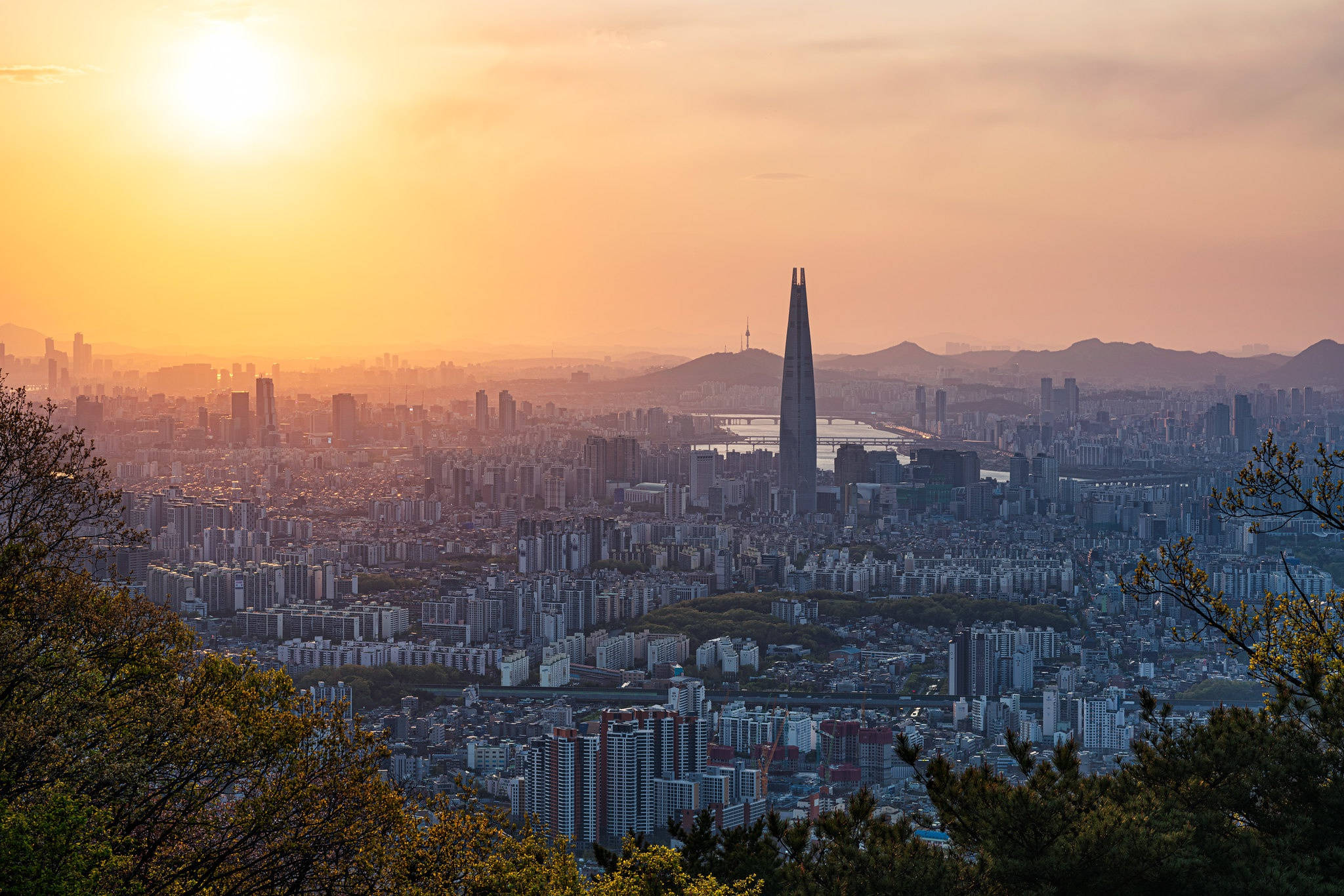 南韓 桌布 壁紙 — 金色的夕陽以溫暖的色調沐浴著首爾廣闊的天際線，樂天世界塔刺穿了朦朧的地平線。