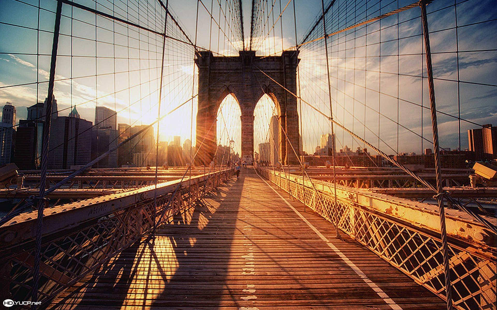 New York 桌布 壁紙 — 金色的夕陽光線穿過布魯克林大橋的拱門，在木製步道上投下長長的影子，背景是城市天際線。