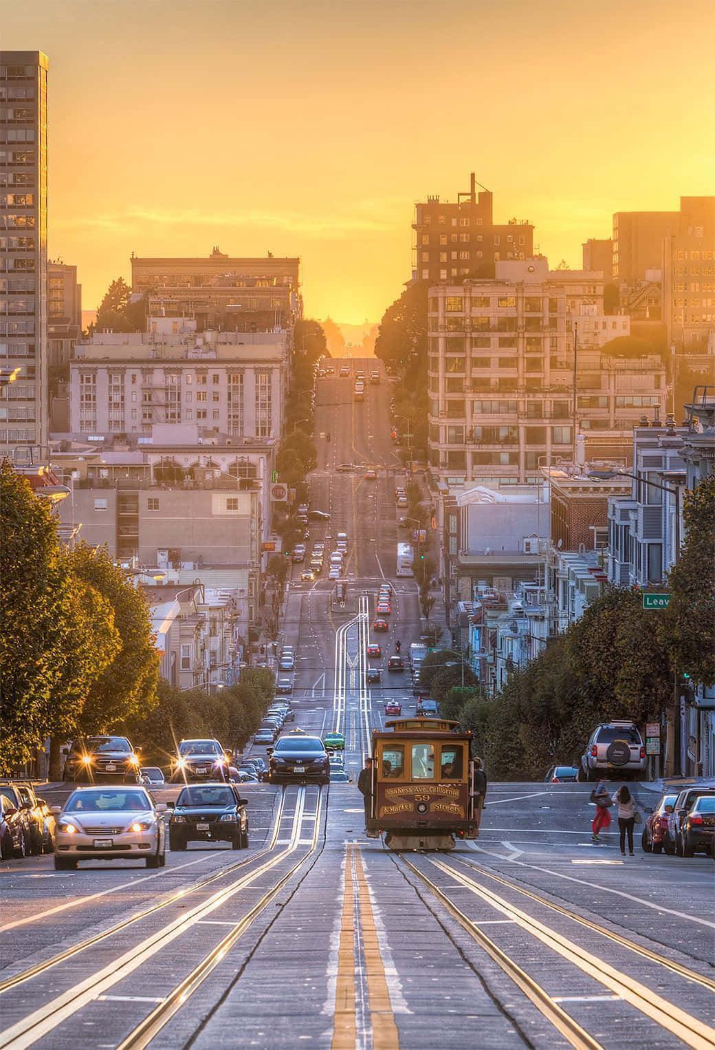 城市 桌布 壁紙 — 金色的夕陽灑在舊金山陡峭的纜車軌道上，一輛復古纜車正沿著兩旁建築物和樹木林立的繁華城市街道上行。
