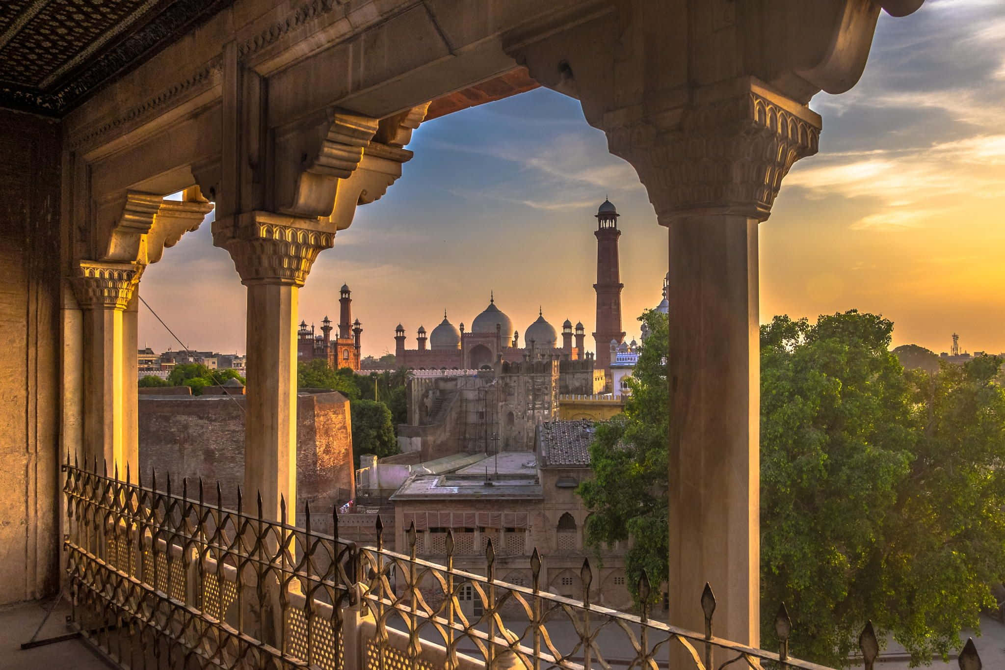 Pakistan 桌布 壁紙 — 金色的夕陽灑落在古老的石拱門和圓頂清真寺建築上，周圍環繞著茂密的綠樹。