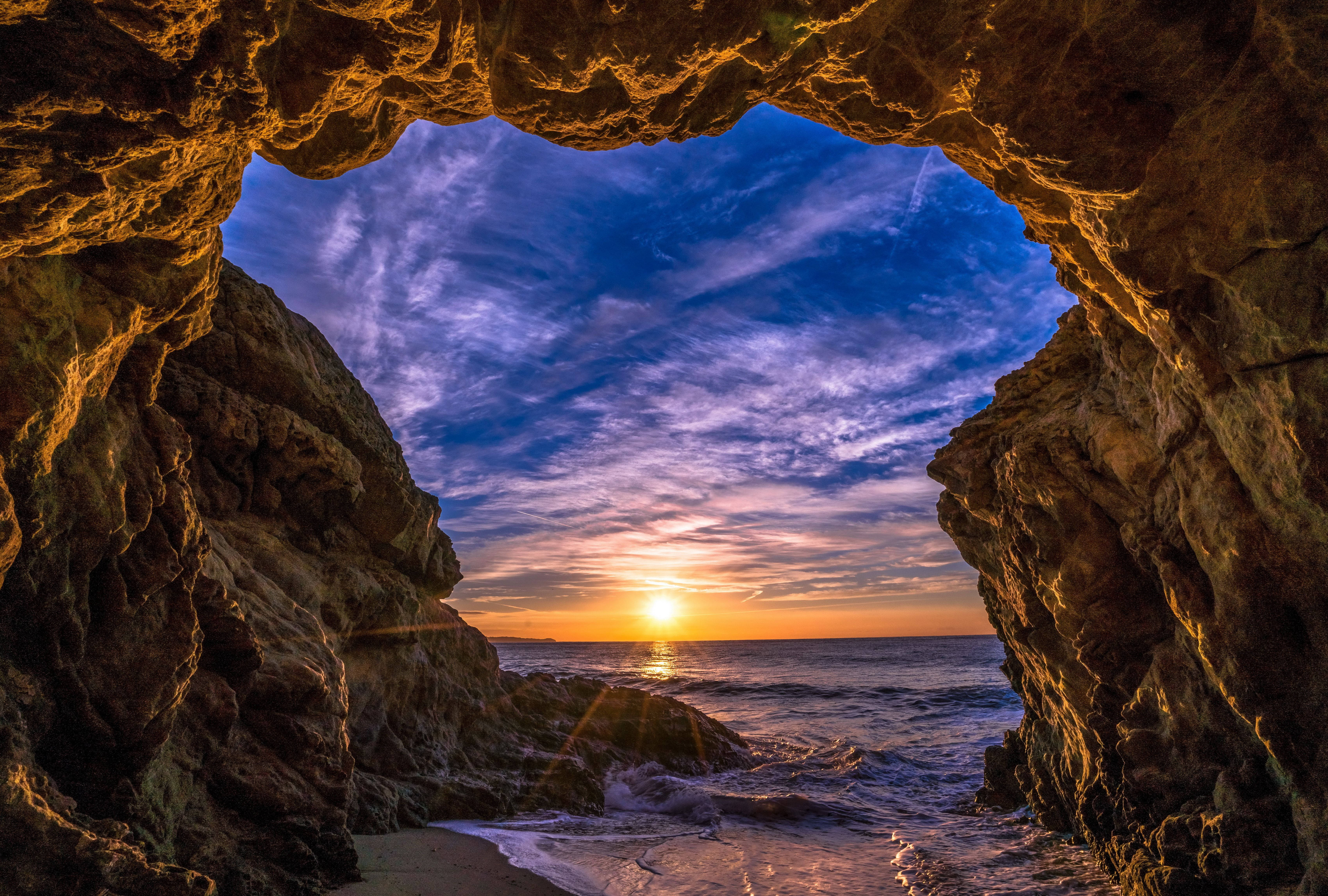 Malibu 桌布 壁紙 — 金色的夕陽透過崎嶇的海蝕洞照射，在翻滾的海浪上方勾勒出充滿活力的紫色和橙色天空。