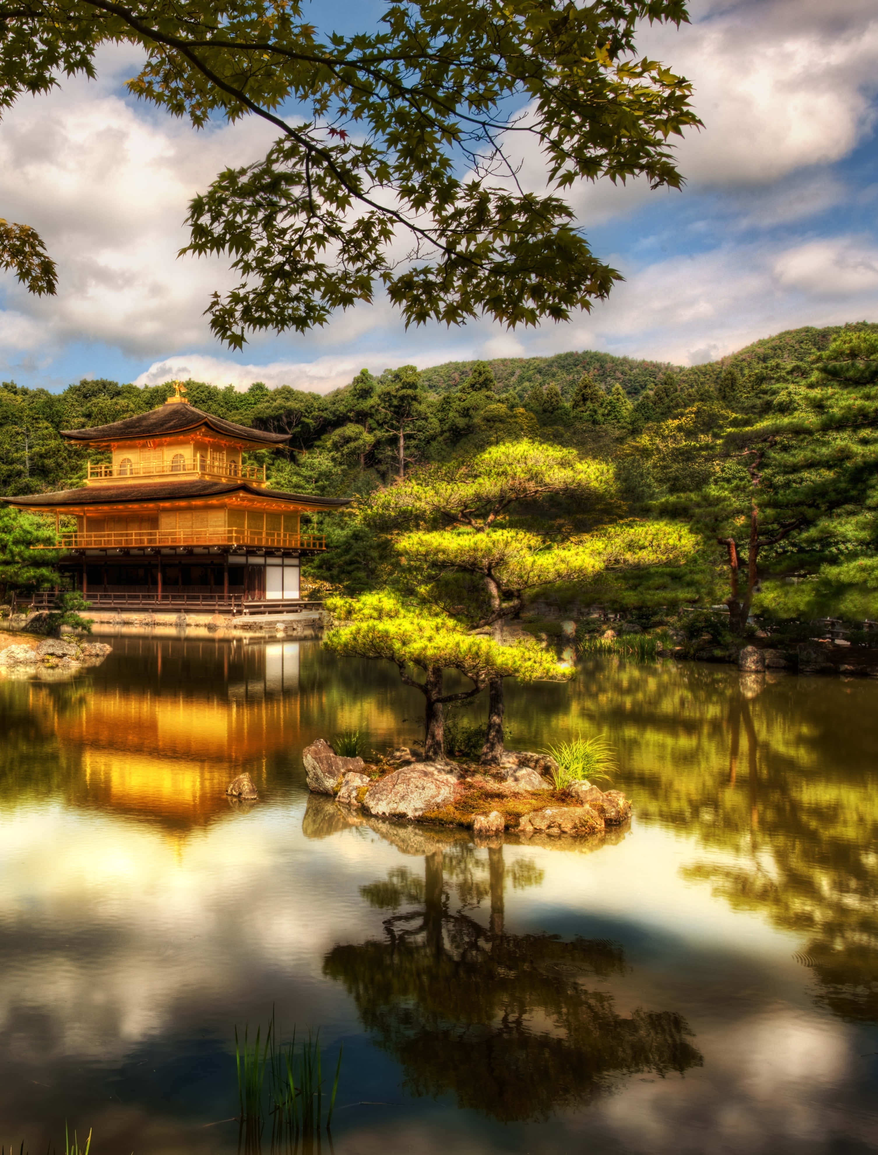 日本 桌布 壁紙 — 金色的金閣寺在部分多雲的天空下，在平靜的水面中倒映，周圍是茂密的綠色樹木。