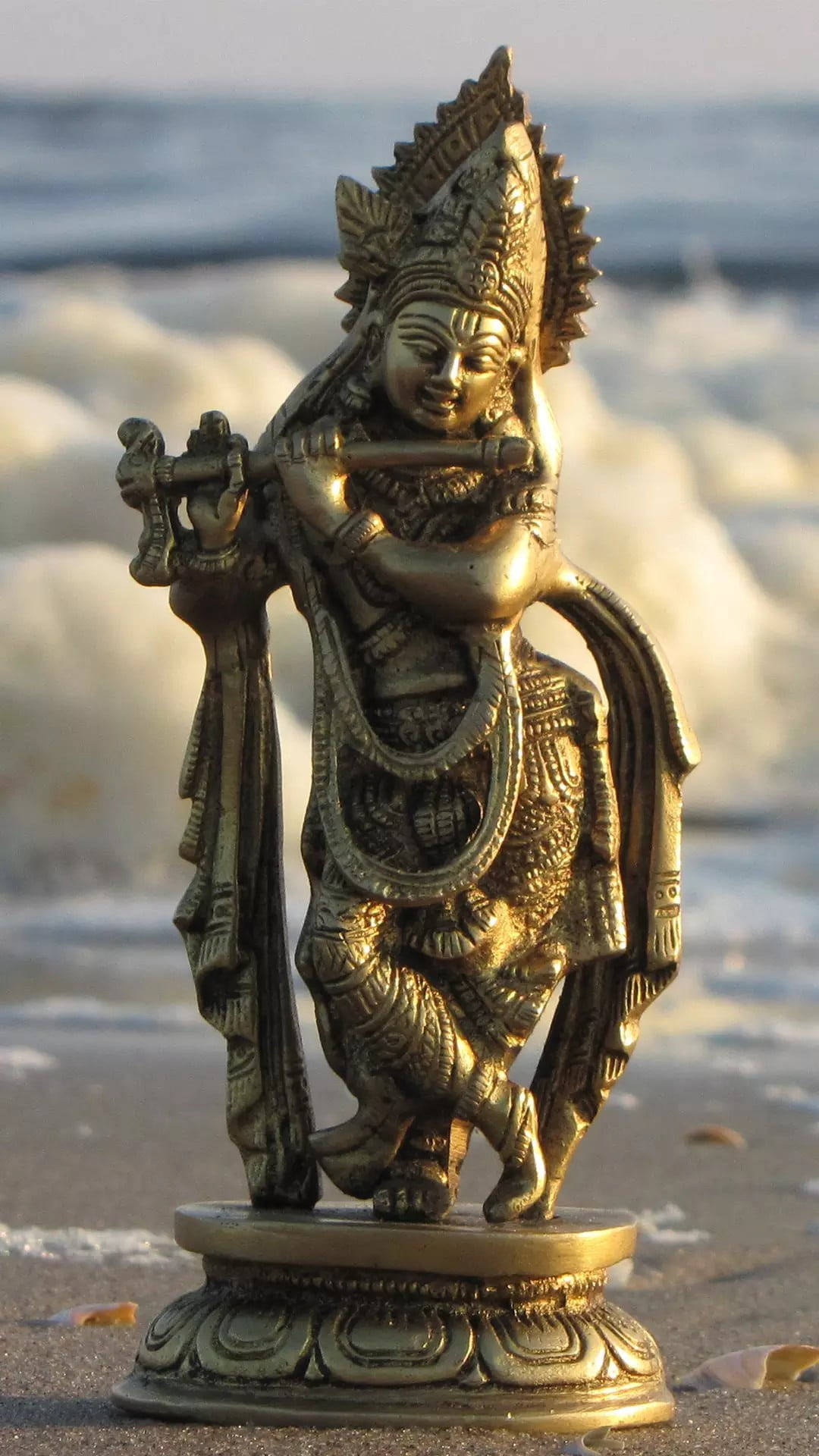 hindu iPhone 桌布 壁紙 — 金色的黃銅克里希納神吹笛雕像矗立在海灘上，背後有海浪拍打。
