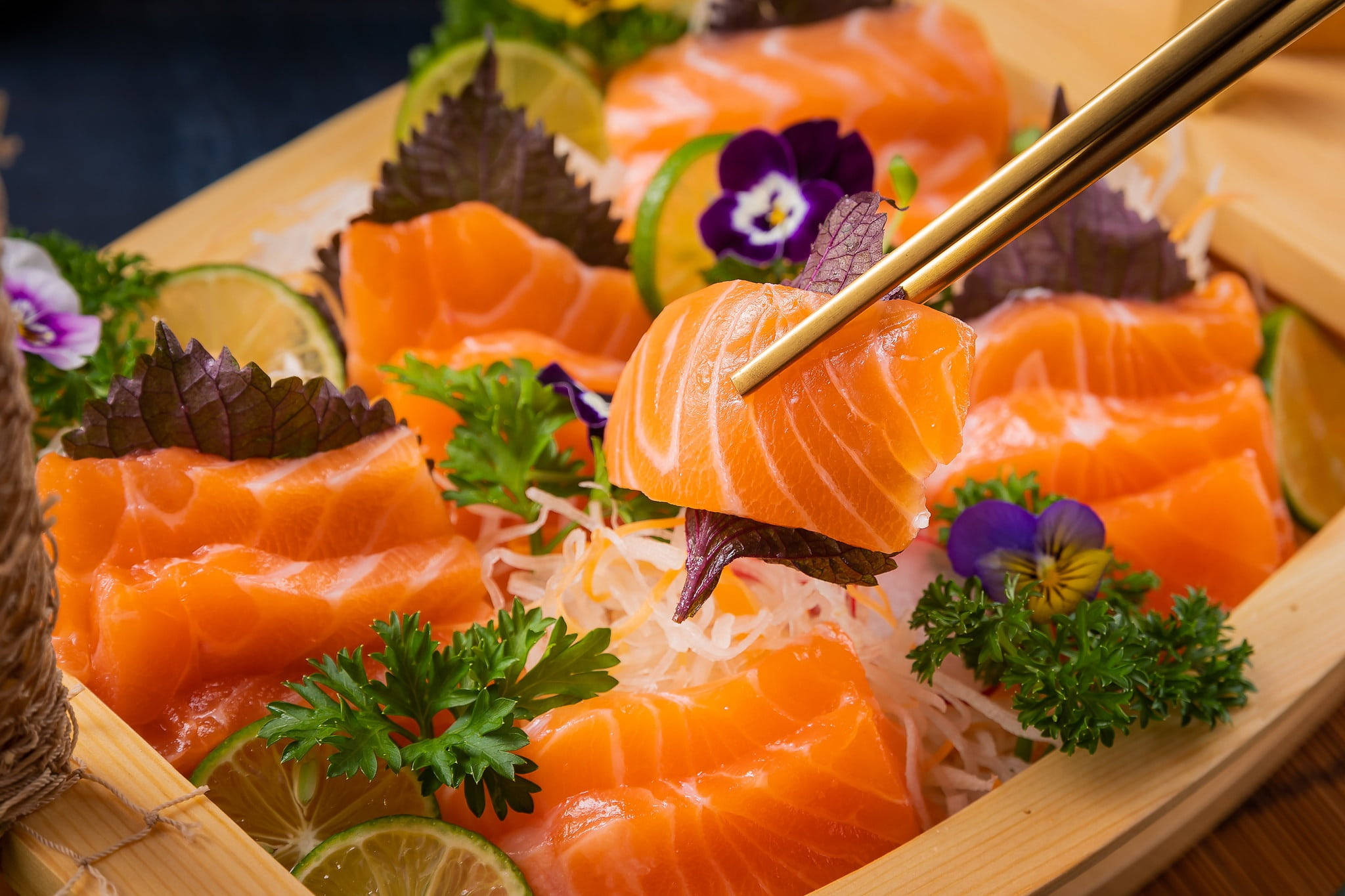 Delicious Food 桌布 壁紙 — 金色筷子將一片鮮豔的橙色鮭魚刺身高高舉起，放在一個飾有紫色花朵、萊姆角和蘿蔔絲的木盤上。