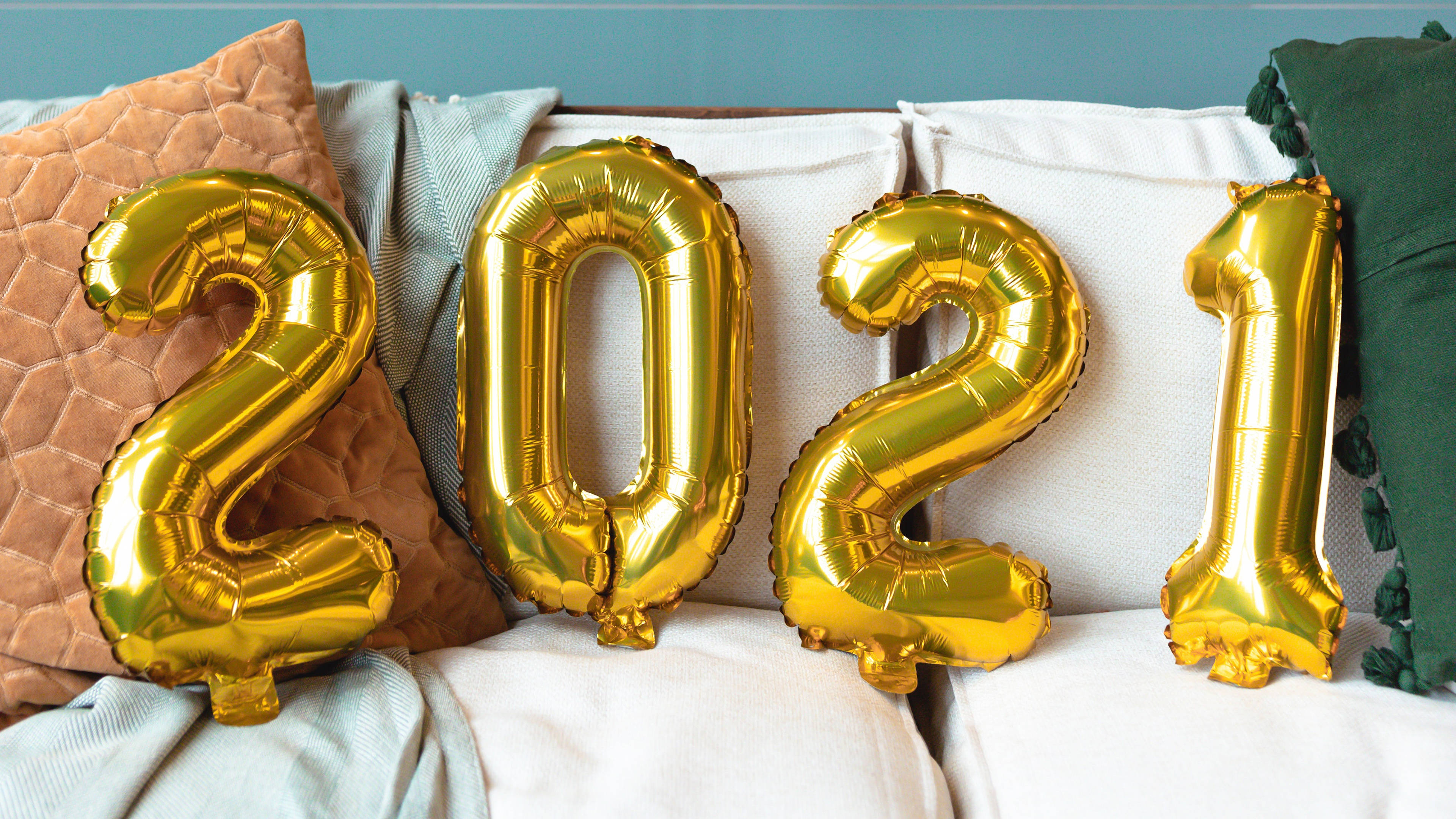 Balloons 桌布 壁紙 — 金色箔氣球拼出2021，放在一張舒適的沙發上，搭配有紋理的靠墊，背景是藍綠色牆壁。