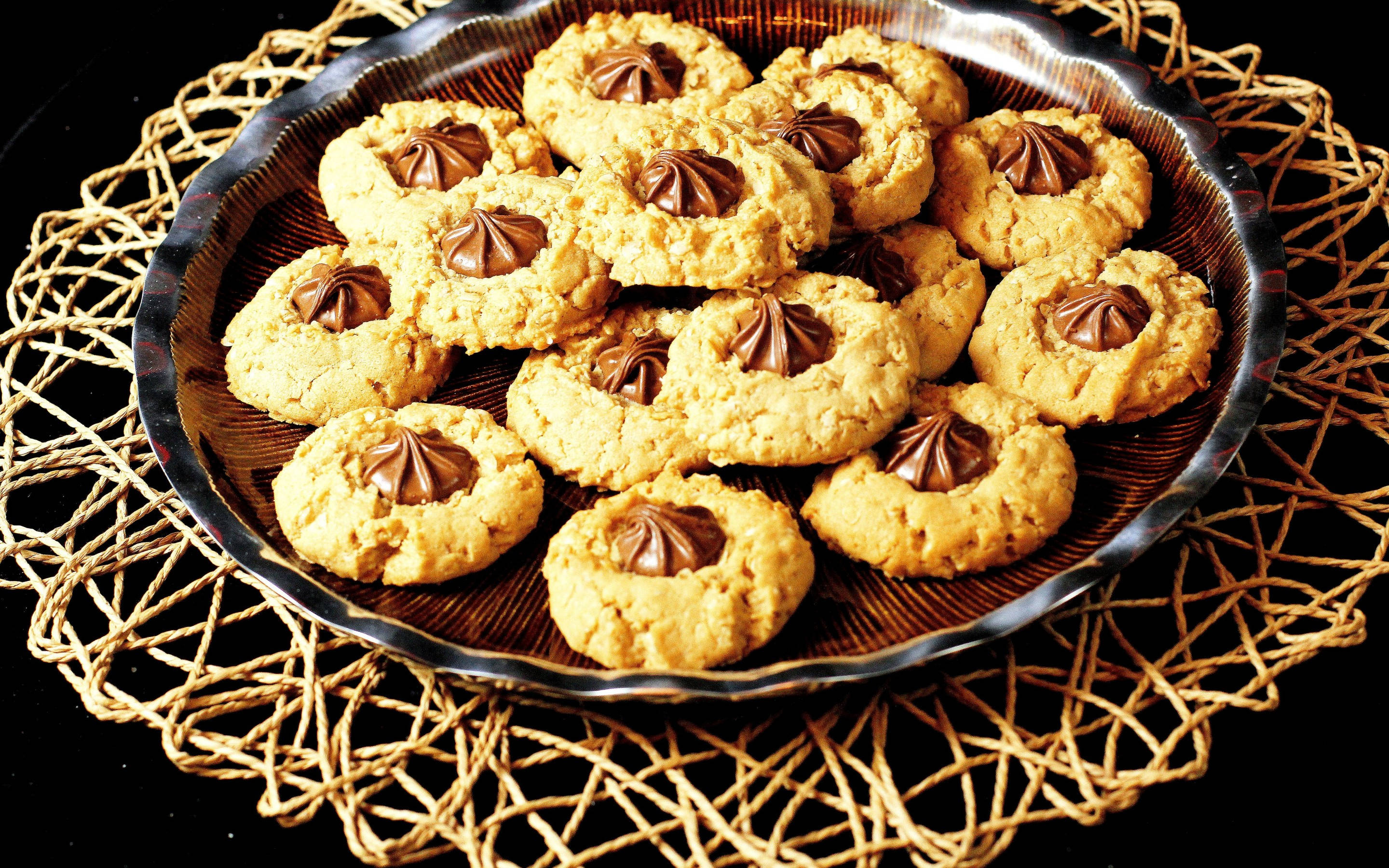 Cookie 桌布 壁紙 — 金色花生醬餅乾，中心有巧克力，堆疊在深色玻璃盤上，置於編織墊上，背景為黑色。