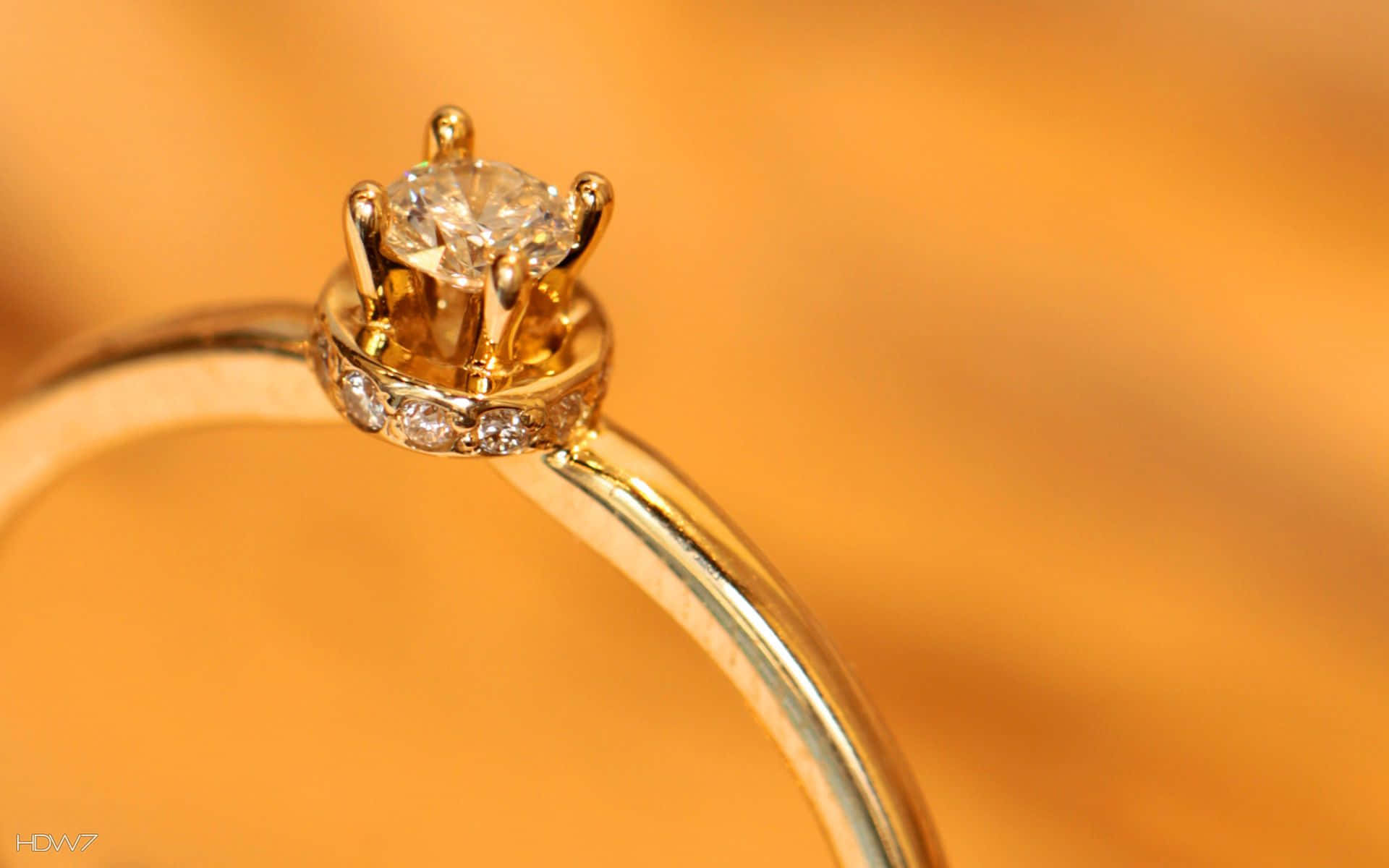 rings 桌布 壁紙 — 金色訂婚戒指的特寫，中央鑲有主鑽和碎鑽光環，背景為溫暖的橙色。