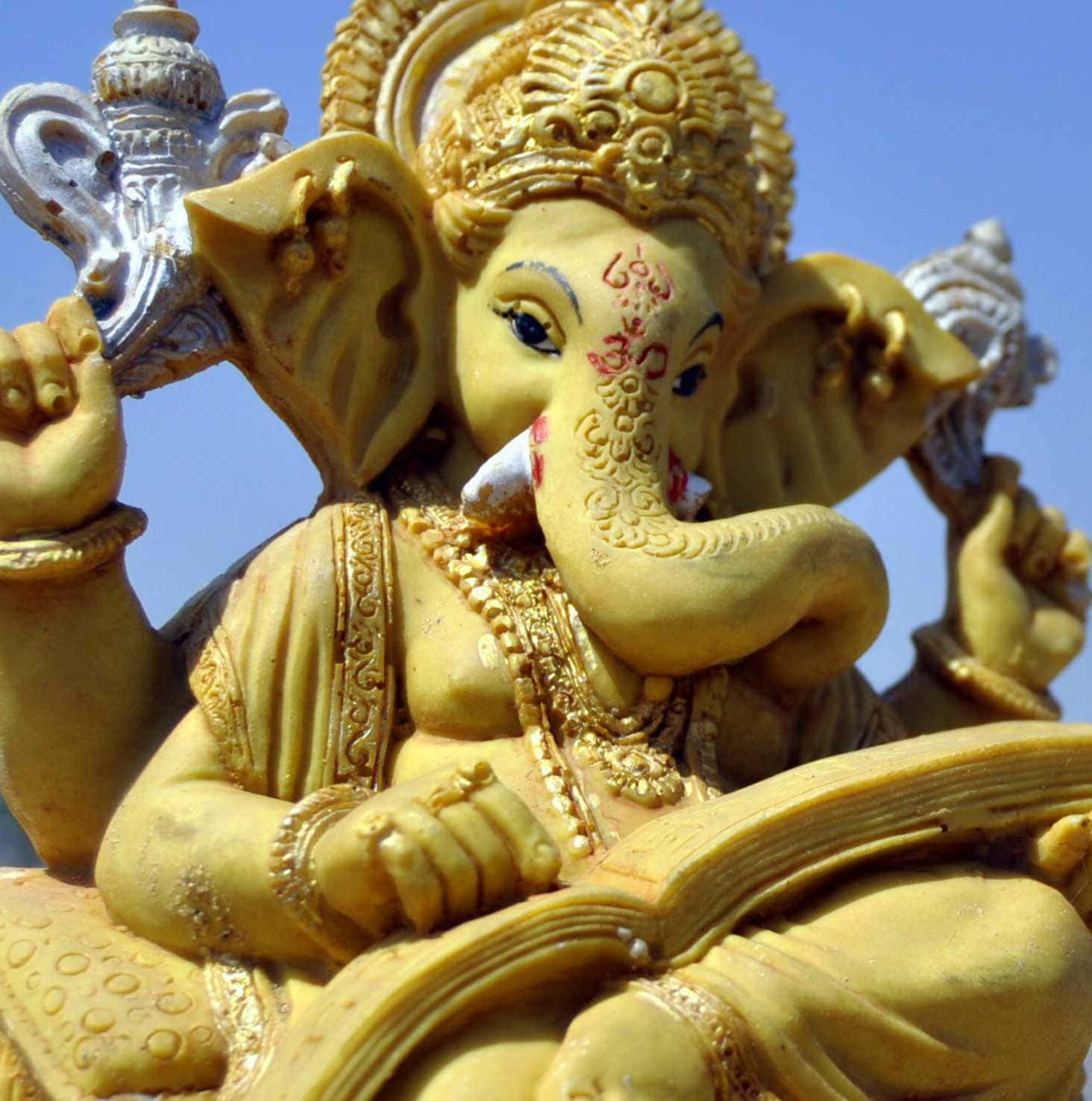 Ganesh 桌布 壁紙 — 金色象神雕像，細節精緻，手持書本和斧頭，背景是清澈的藍天。