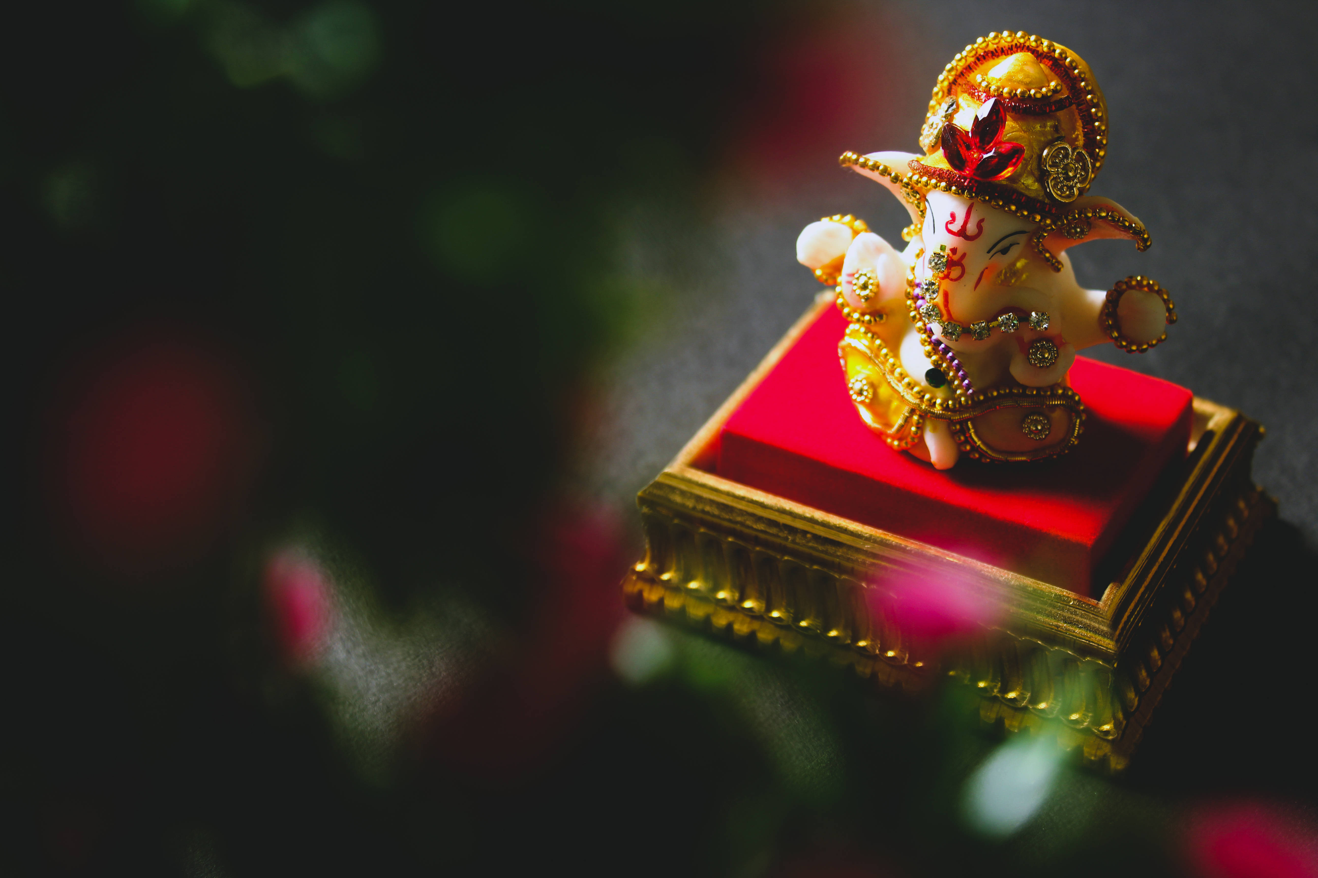 神 桌布 壁紙 — 金色象神雕像威嚴地坐在紅色軟墊底座上，背景是模糊的深色花卉。