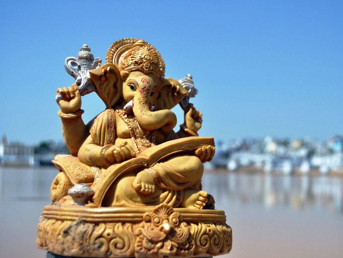 Hindu God 桌布 壁紙 — 金色象神雕像威嚴地坐在華麗的底座上，手持書本和斧頭，背景是模糊的河流和城市景觀，在清澈的藍天下。