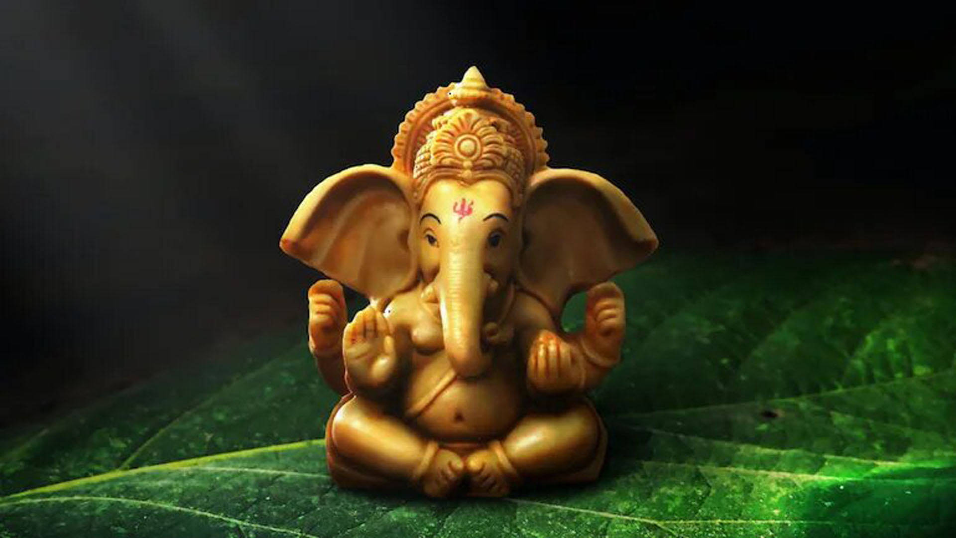 Hindu God 桌布 壁紙 — 金色象神雕像安詳地坐在深色背景的大綠葉上。