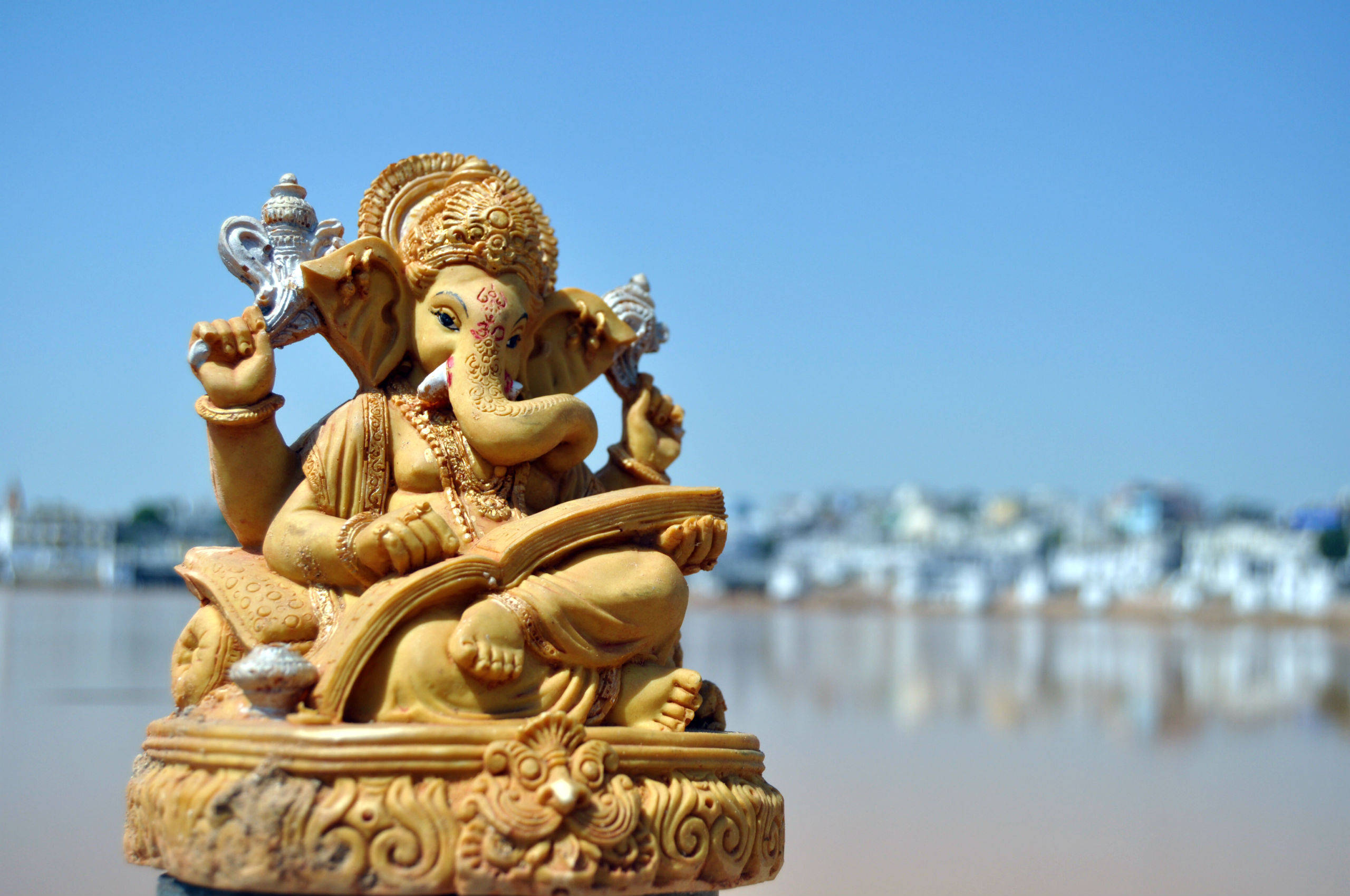 Ganesh 桌布 壁紙 — 金色象神雕像靜靜地坐在河邊，模糊的城市景觀在清澈的藍天下。