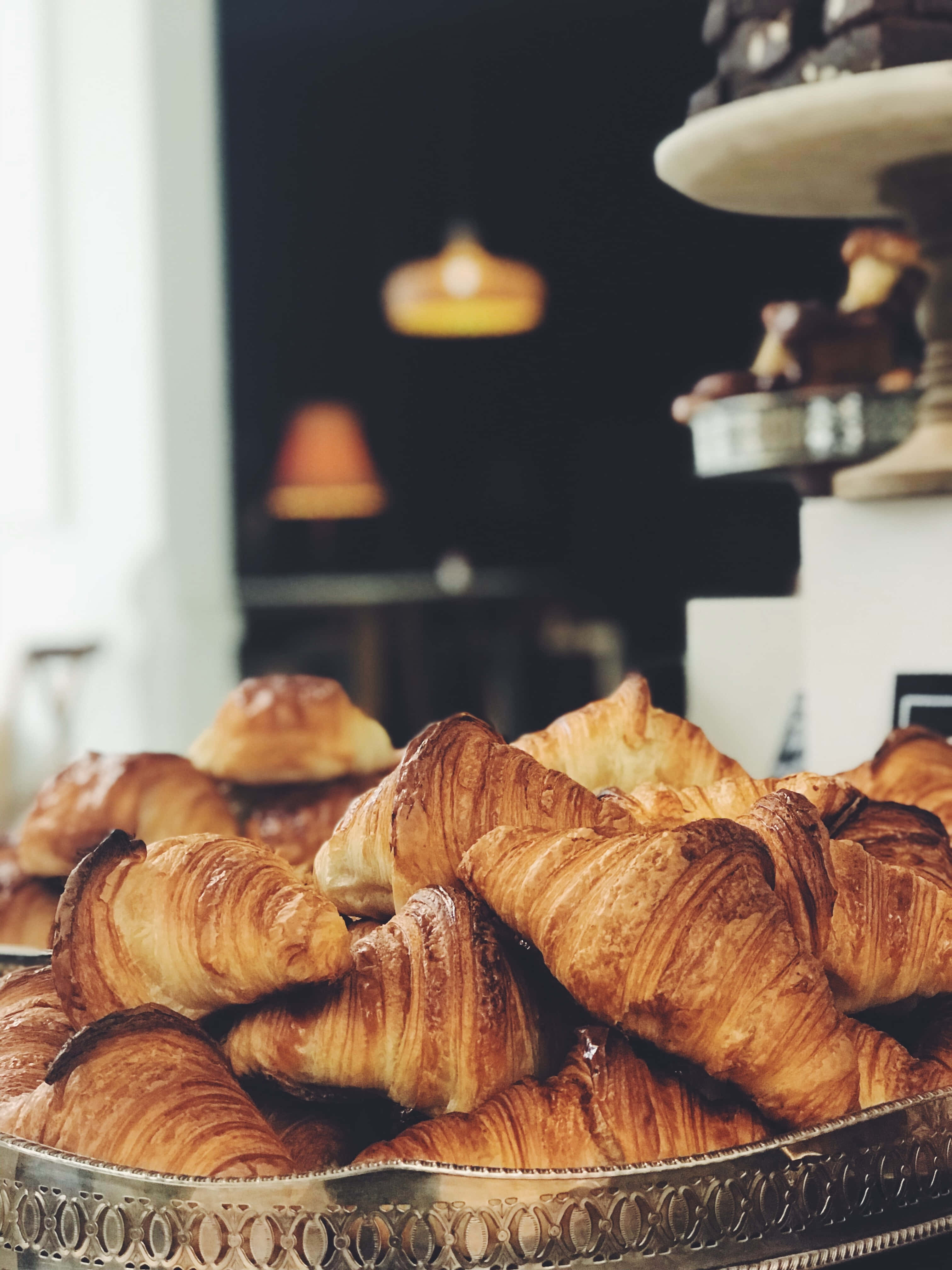 Bakery 桌布 壁紙 — 金色酥脆可頌高高堆放在華麗的銀托盤上，背景中溫暖的咖啡館燈光柔和地閃爍的特寫。