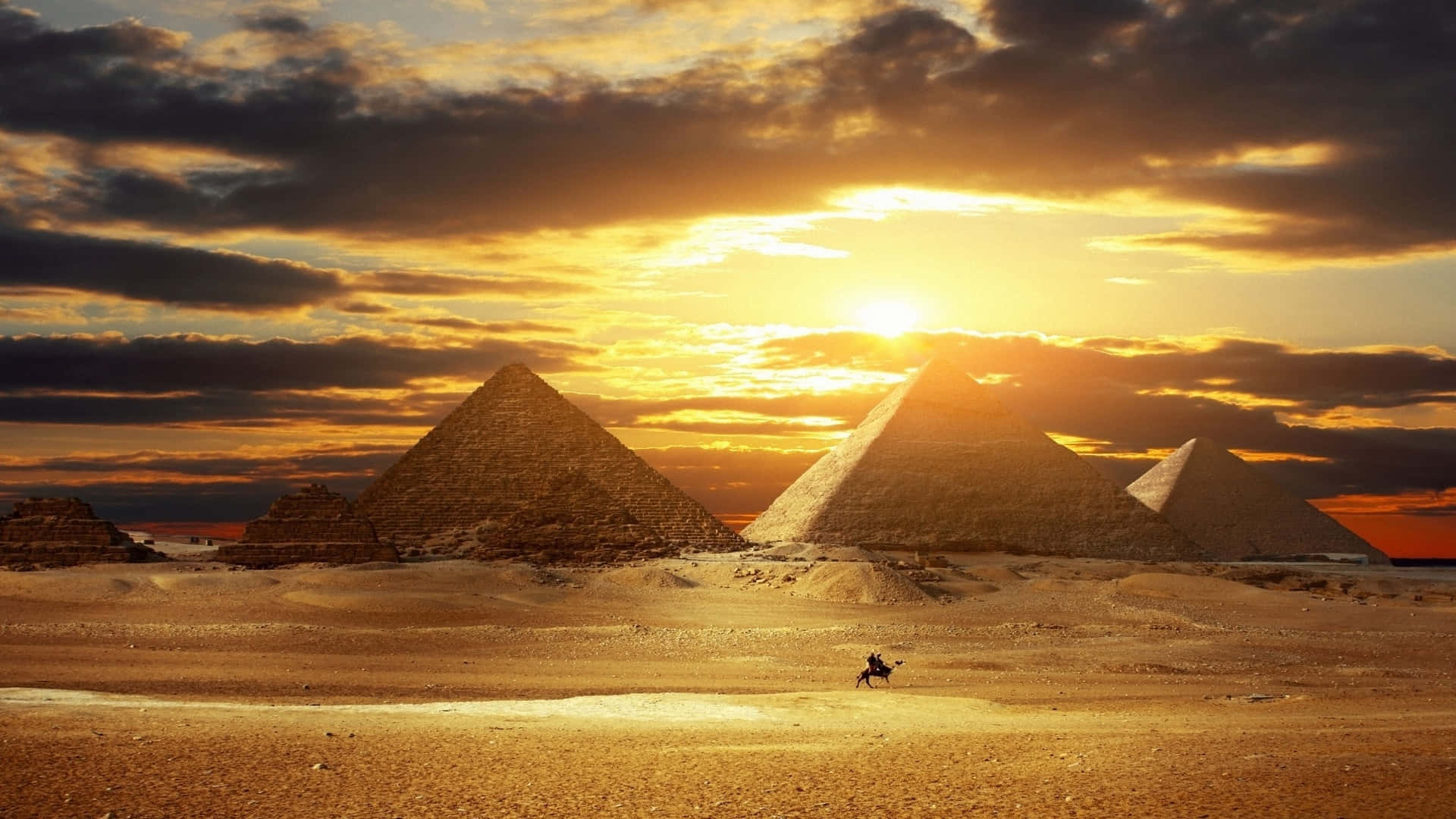 egyptian 桌布 壁紙 — 金色金字塔從沙漠沙地升起，一名孤獨的騎手騎著駱駝在熾熱的夕陽天空下穿越沙丘。