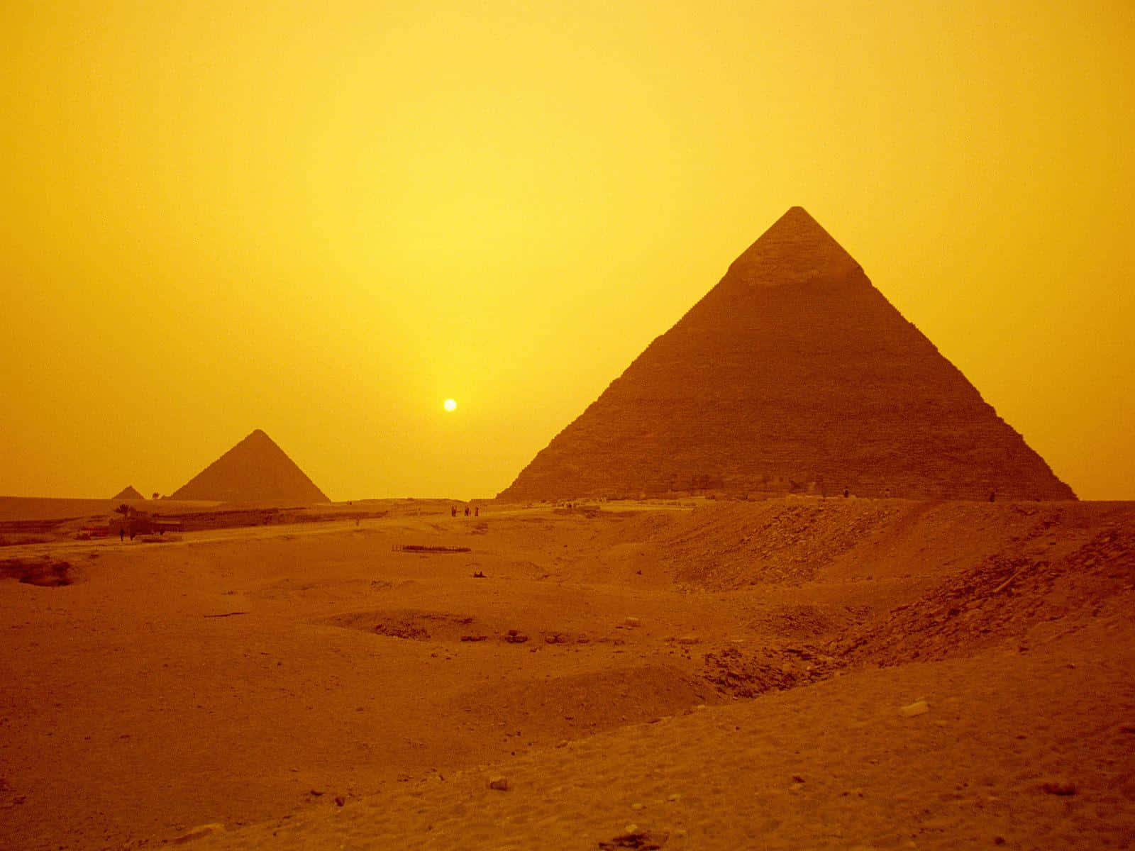 Egypt 桌布 壁紙 — 金色金字塔從沙漠沙地升起，在朦朧的橙色日落天空下，明亮的太陽低掛在地平線上。