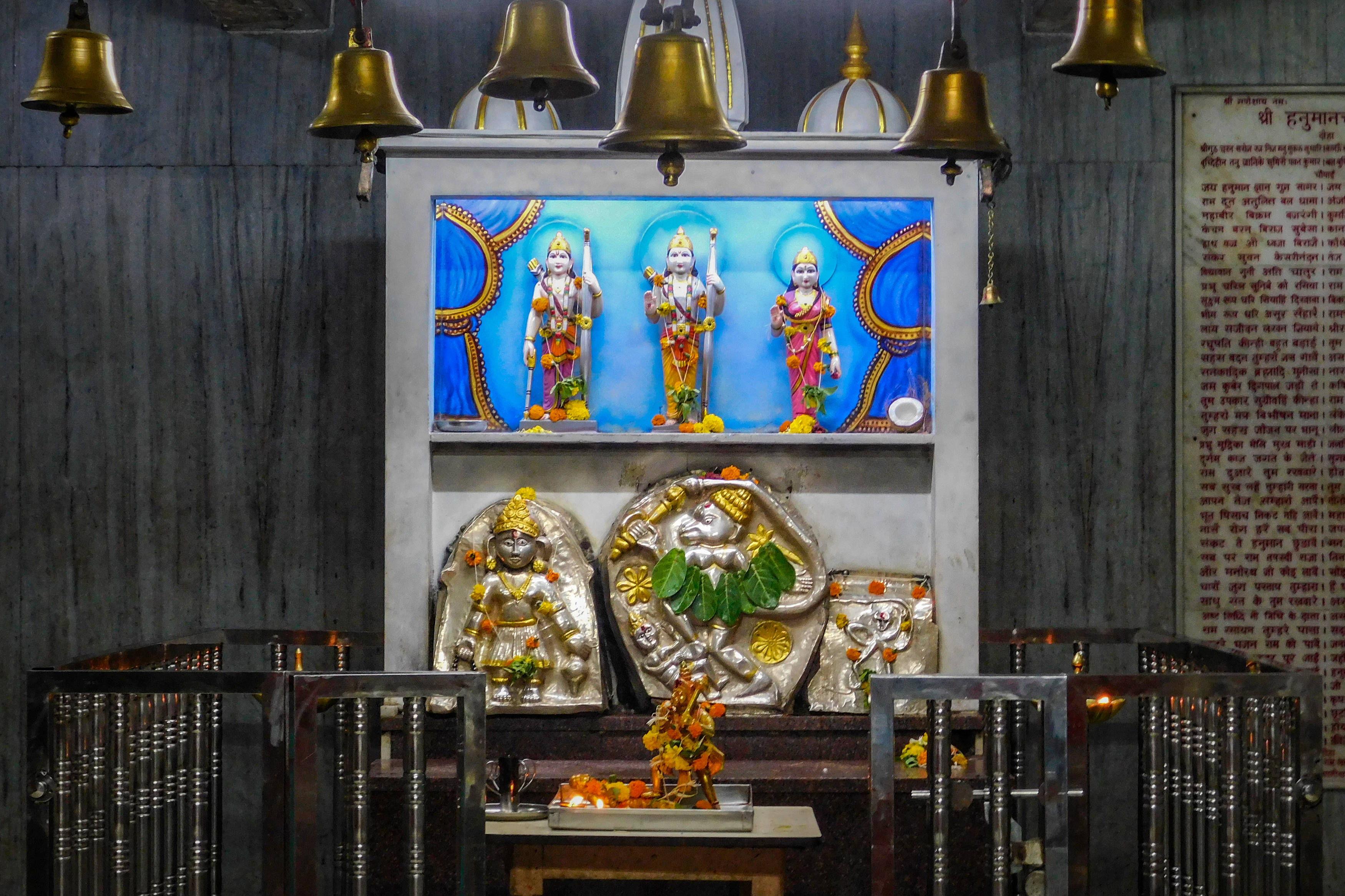 Hindu Temple 桌布 壁紙 — 金色鐘鈴懸掛在色彩鮮豔的印度教神像上方，位於燈光昏暗的寺廟神龕中，下方有銀色神像和供品。