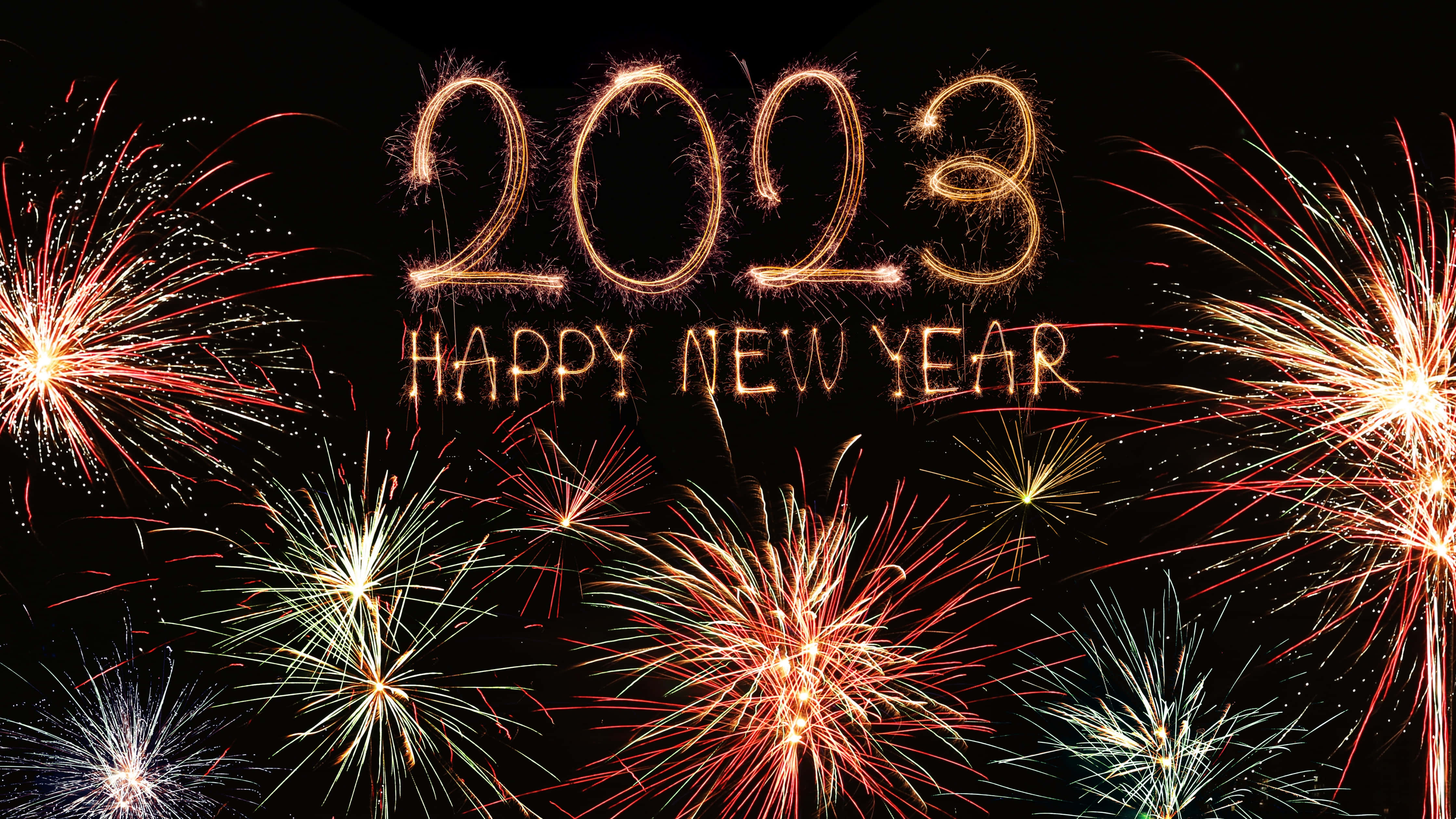 Fireworks 桌布 壁紙 — 金色閃亮的「2023 HAPPY NEW YEAR」文字在佈滿爆裂的紅色、白色和綠色煙火的黑色天空中發光。