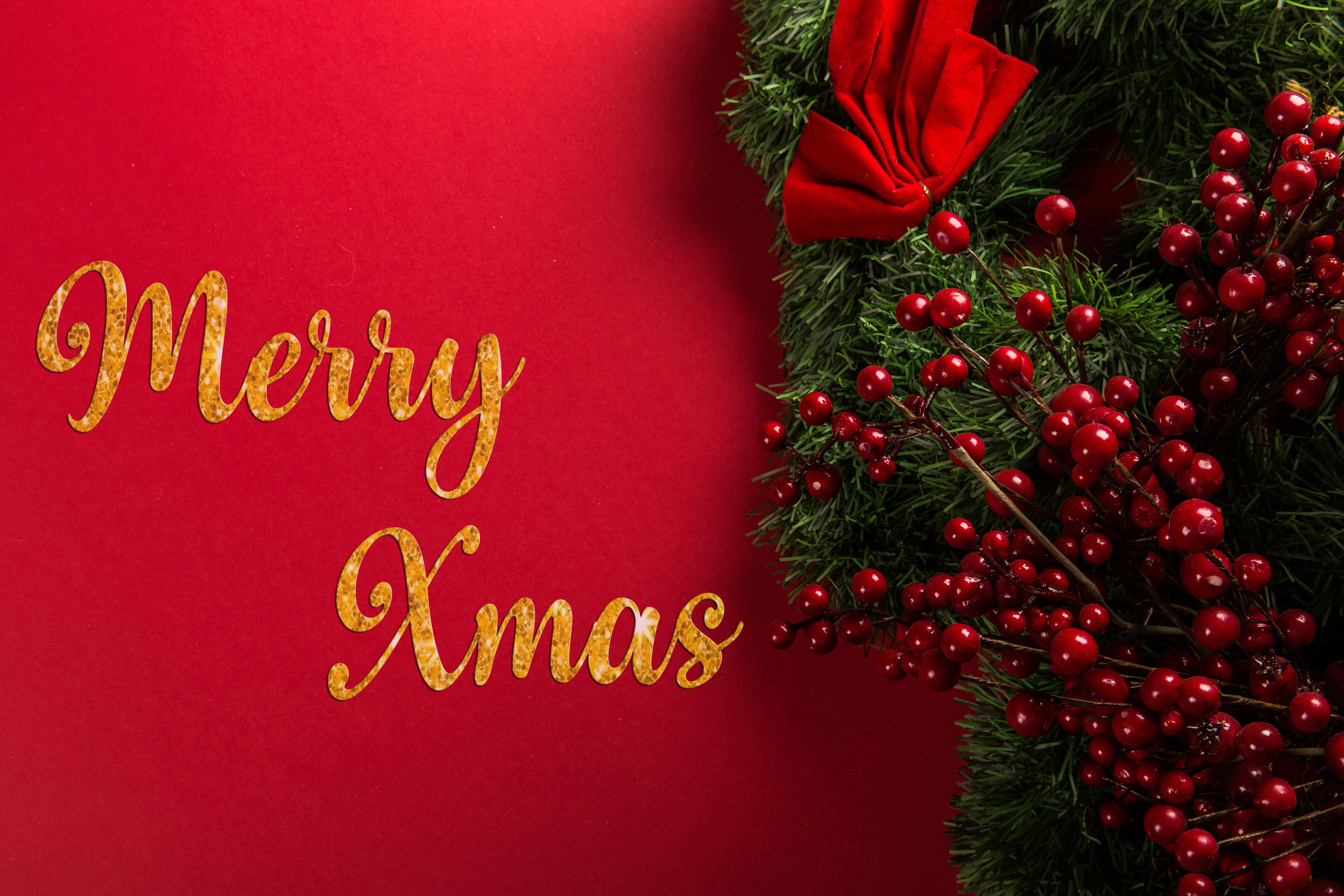 節日 桌布 壁紙 — 金色閃閃發光的Merry Xmas文字在鮮豔的紅色背景上，旁邊是帶有紅色漿果和蝴蝶結的節日綠色花環。