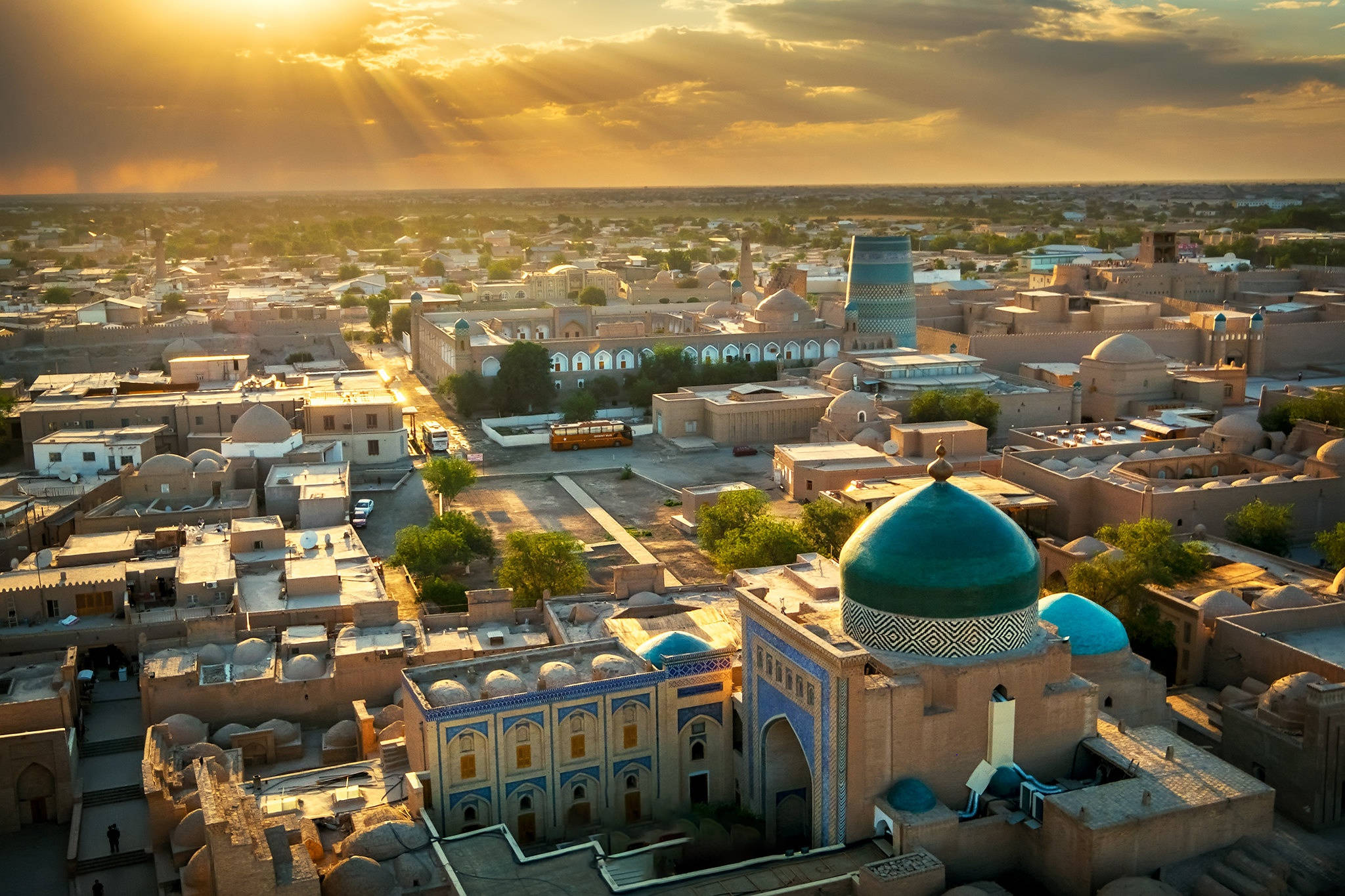 Uzbekistan 桌布 壁紙 — 金色陽光穿透雲層，照耀著擁有綠松色圓頂和大地色建築的歷史城市景觀。