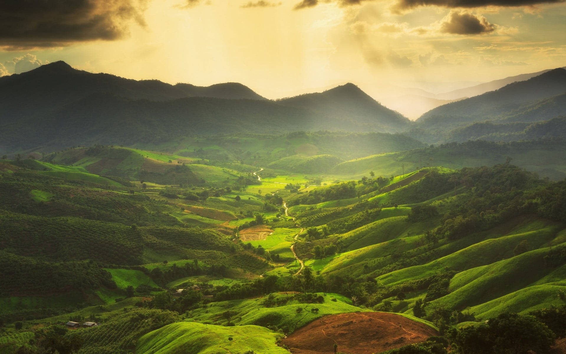 Green Hill 桌布 壁紙 — 金色陽光穿過雲層，照亮了寧靜山谷中起伏的綠色山丘和蜿蜒小徑。