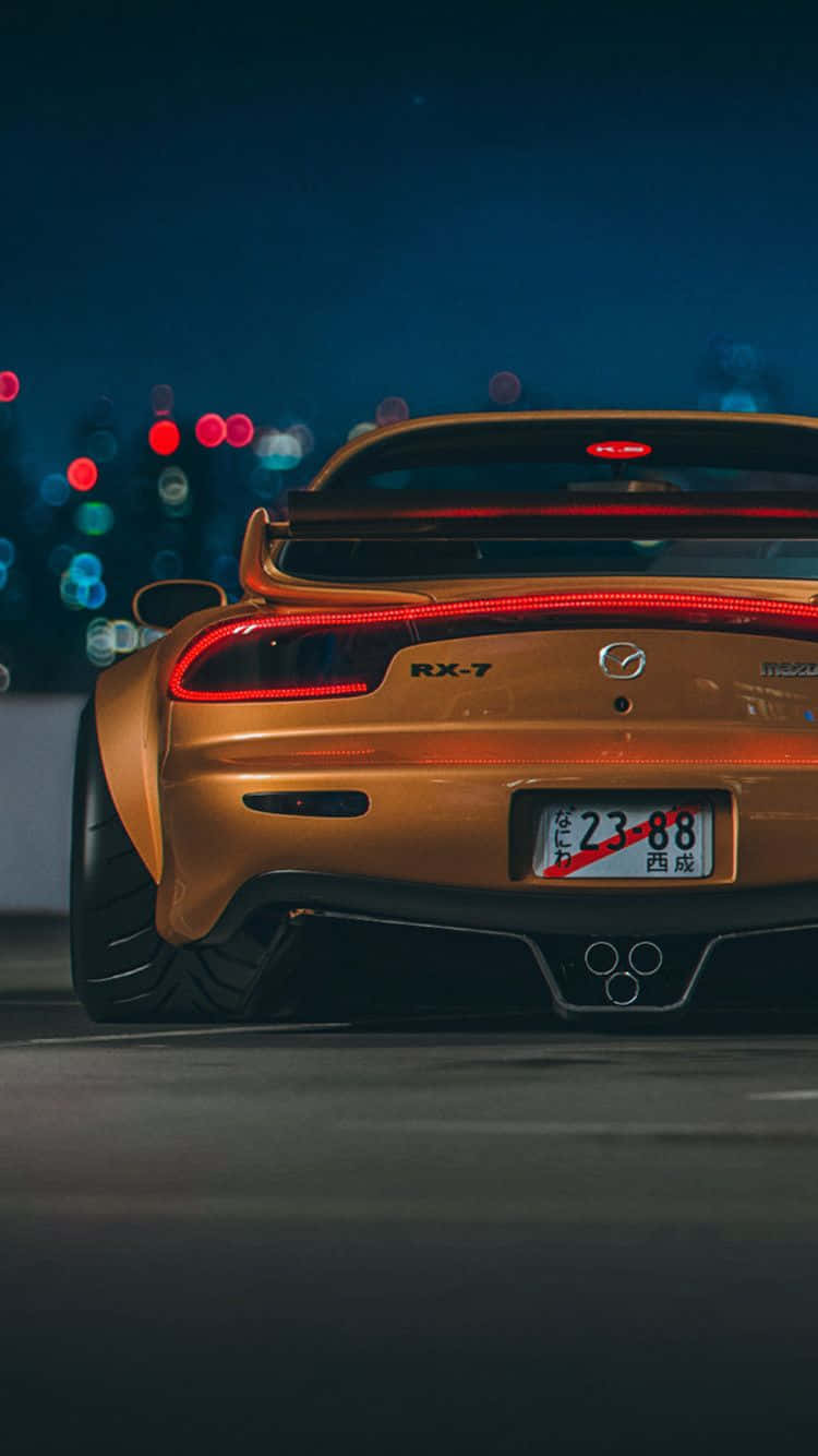 Mazda 桌布 壁紙 — 金色馬自達RX-7跑車，紅色尾燈發光，夜晚停泊在模糊的城市景觀前。