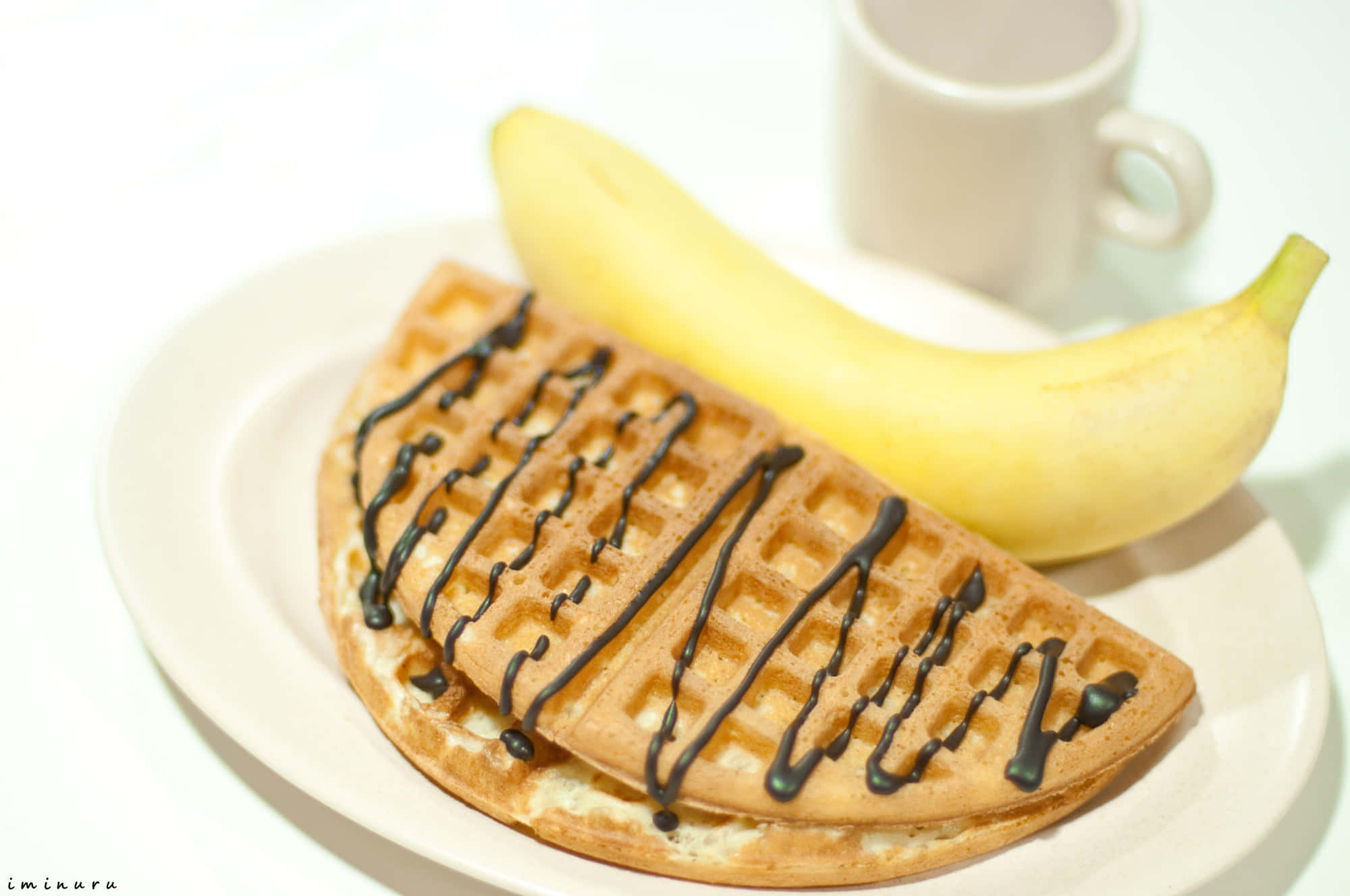 Waffle 桌布 壁紙 — 金色鬆餅淋上巧克力醬，旁邊放著一根熟香蕉，放在白色盤子上，背景有杯模糊的杯子。