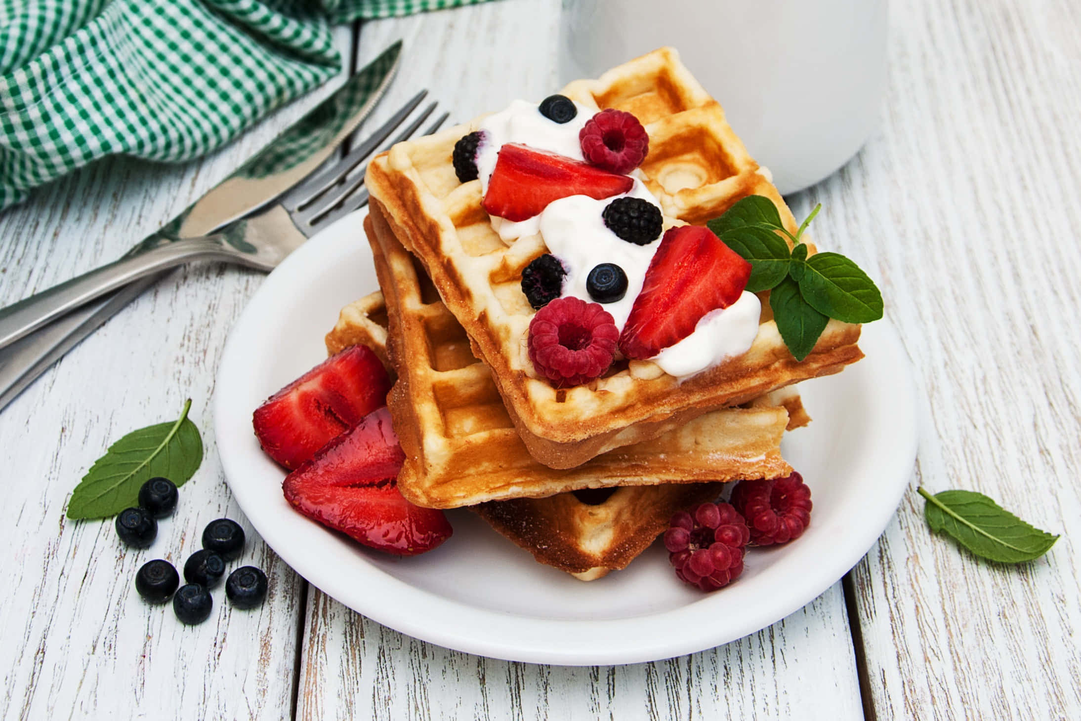 Waffle 桌布 壁紙 — 金色鬆餅高高疊起，配上新鮮莓果、鮮奶油和薄荷，放在質樸的白色木桌上。
