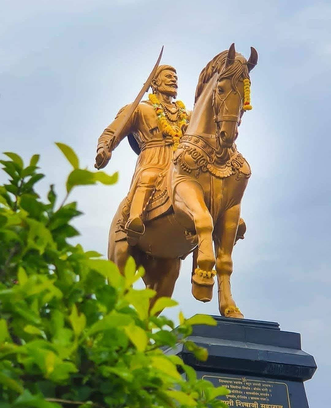 Indian 桌布 壁紙 — 金色鬍鬚戰士雕像，騎在後腿站立的馬上，馬匹飾有花環，背景是淡色天空，由綠葉框住。